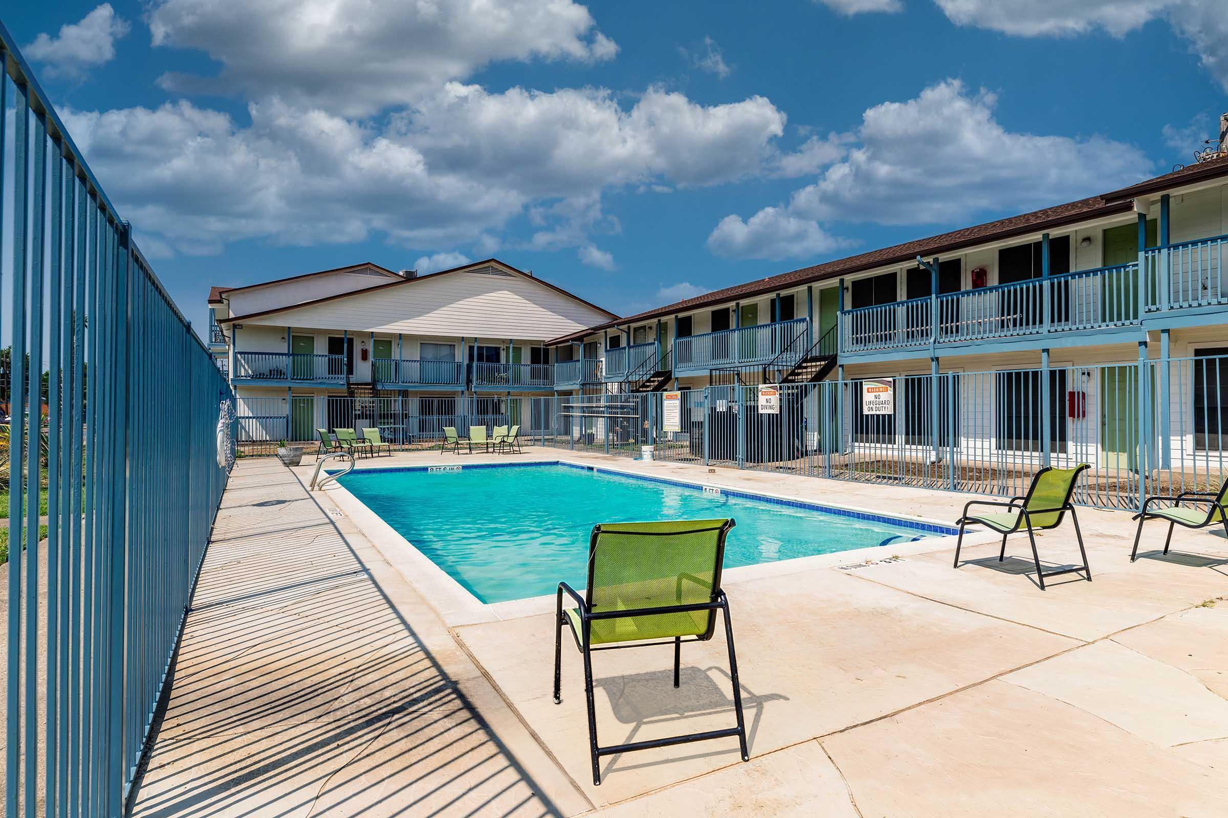 A sunny day at a poolside of a motel, featuring a clear blue swimming pool surrounded by lounge chairs. The two-story building in the background has balconies with green railings and multiple rooms. A fenced area offers privacy, and fluffy clouds dot the blue sky above.