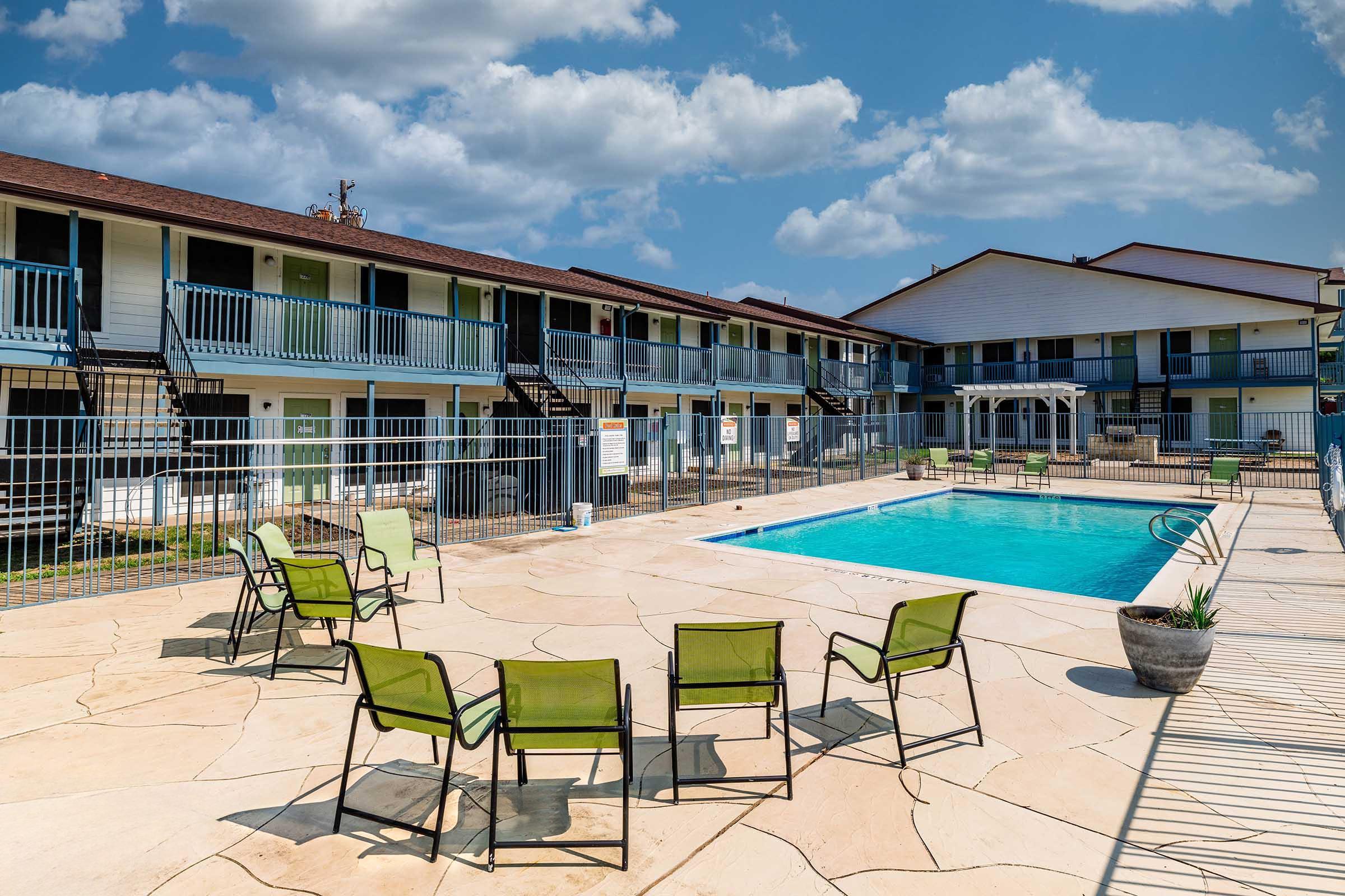 A bright outdoor pool area surrounded by lounge chairs, with a two-story motel-style building in the background. The pool is enclosed by a fence, and there are partly cloudy skies above. A few potted plants are placed around the pool deck, creating a welcoming atmosphere.