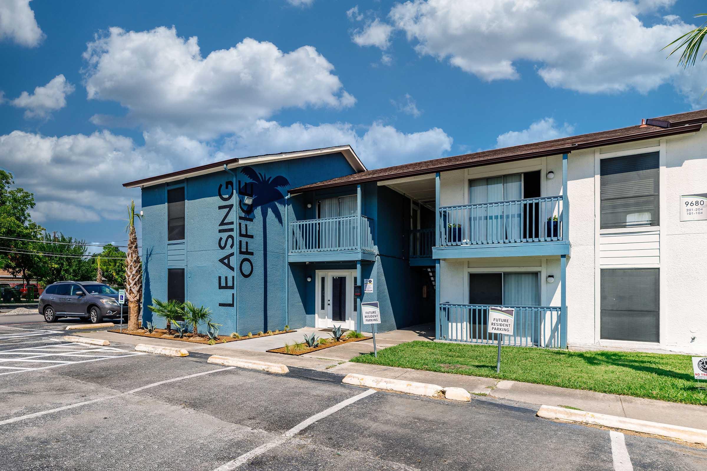 Leasing office of an apartment complex with a blue exterior, palm tree decoration, and multiple balconies. The area features green lawn and parking spaces in front, under a partly cloudy sky.