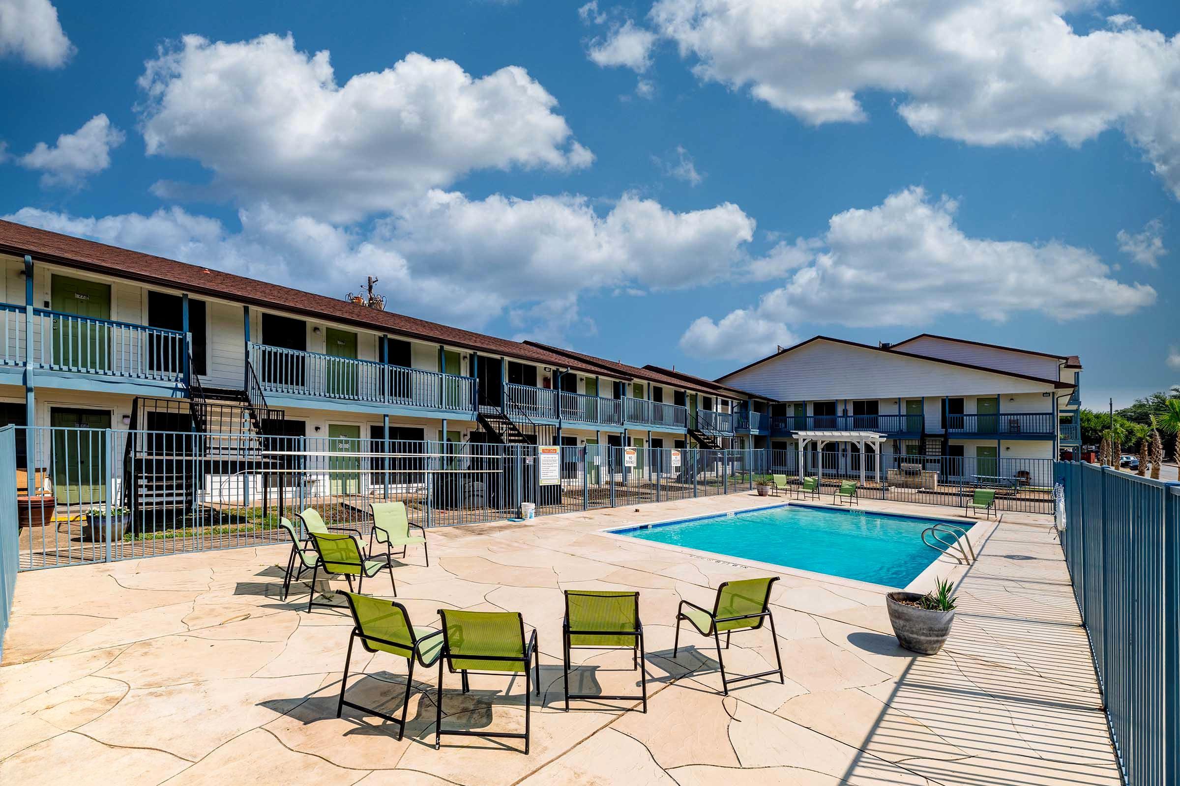A sunny outdoor area featuring a swimming pool surrounded by lounge chairs and a fenced patio. In the background, there are two-story buildings with balconies and stairs. The sky is partly cloudy, enhancing the bright and inviting atmosphere of the scene.