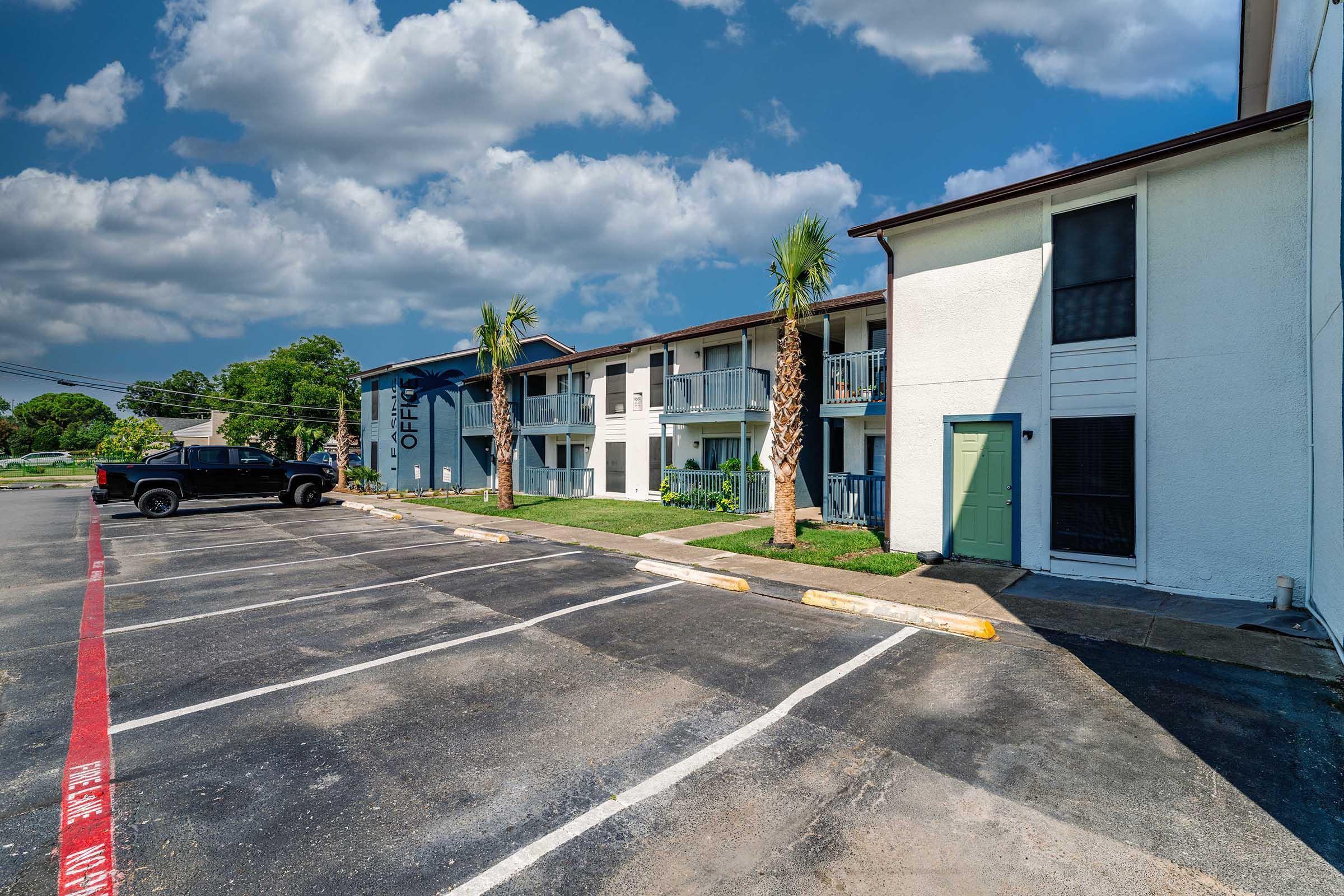 A view of a multi-story motel with palm trees in front, featuring a mix of blue and white exterior walls. A black pickup truck is parked in a partially empty parking lot, with a recently painted red line visible on the pavement. The sky is mostly sunny with scattered clouds.