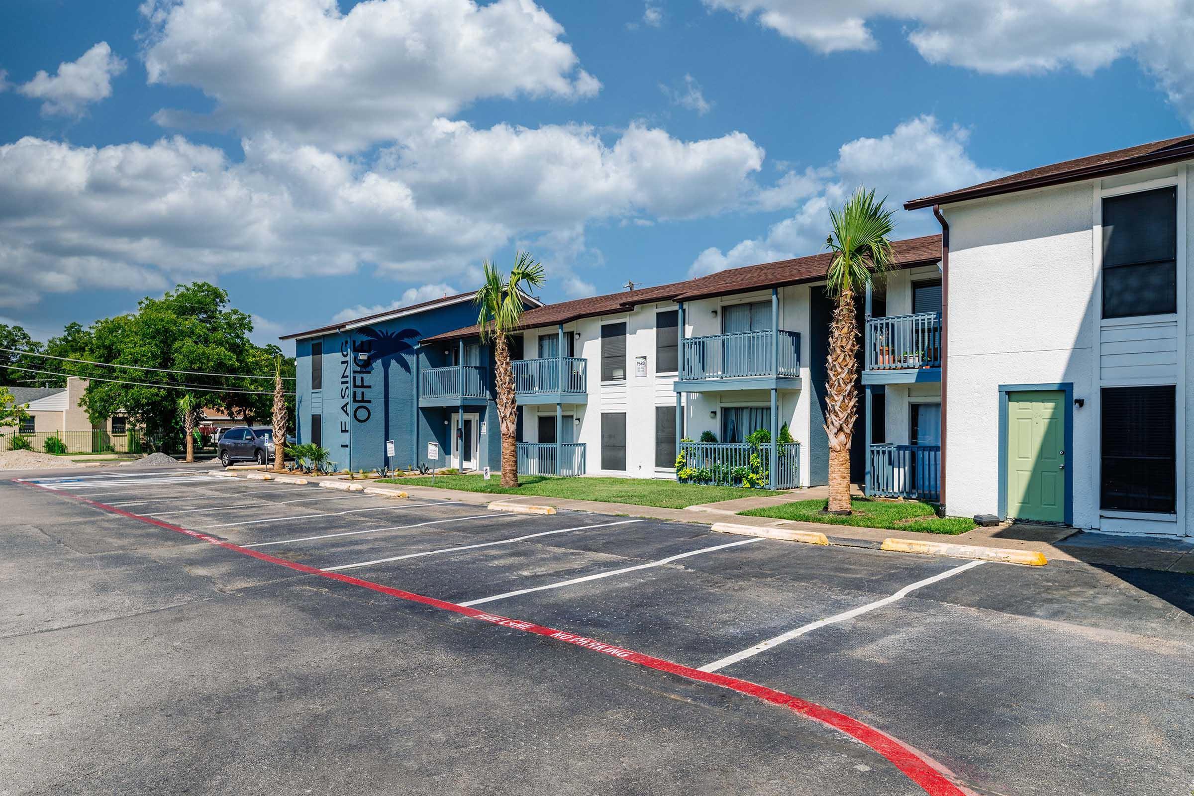 View of a residential apartment complex with a blue sky and fluffy white clouds. The building features palm trees and a parking area with clearly marked spaces. On the side of the building, there's a sign indicating "Office" along with a green door, creating a welcoming atmosphere.