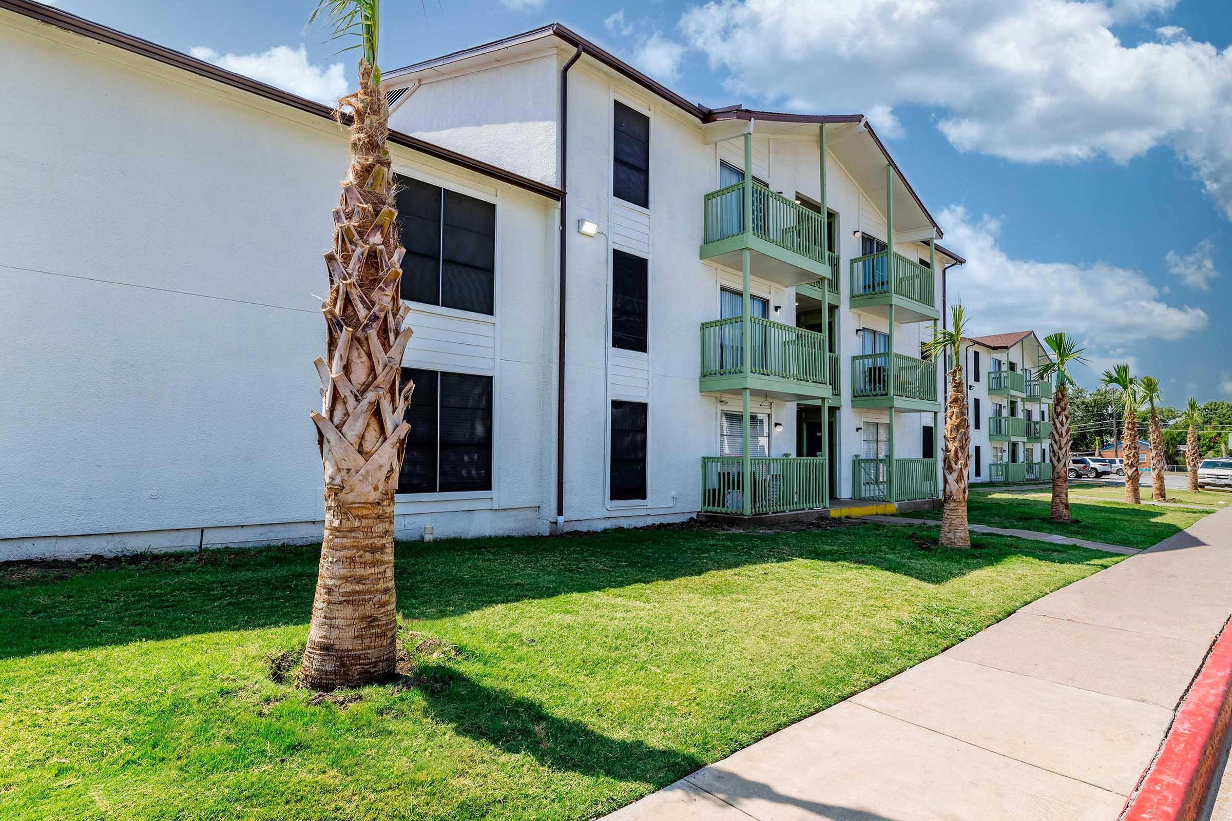 A modern multi-story residential building with white exterior walls and green balconies. Palm trees line the grassy area in front, and a concrete sidewalk runs alongside the property under a partly cloudy sky. The scene conveys a warm, sunny atmosphere.