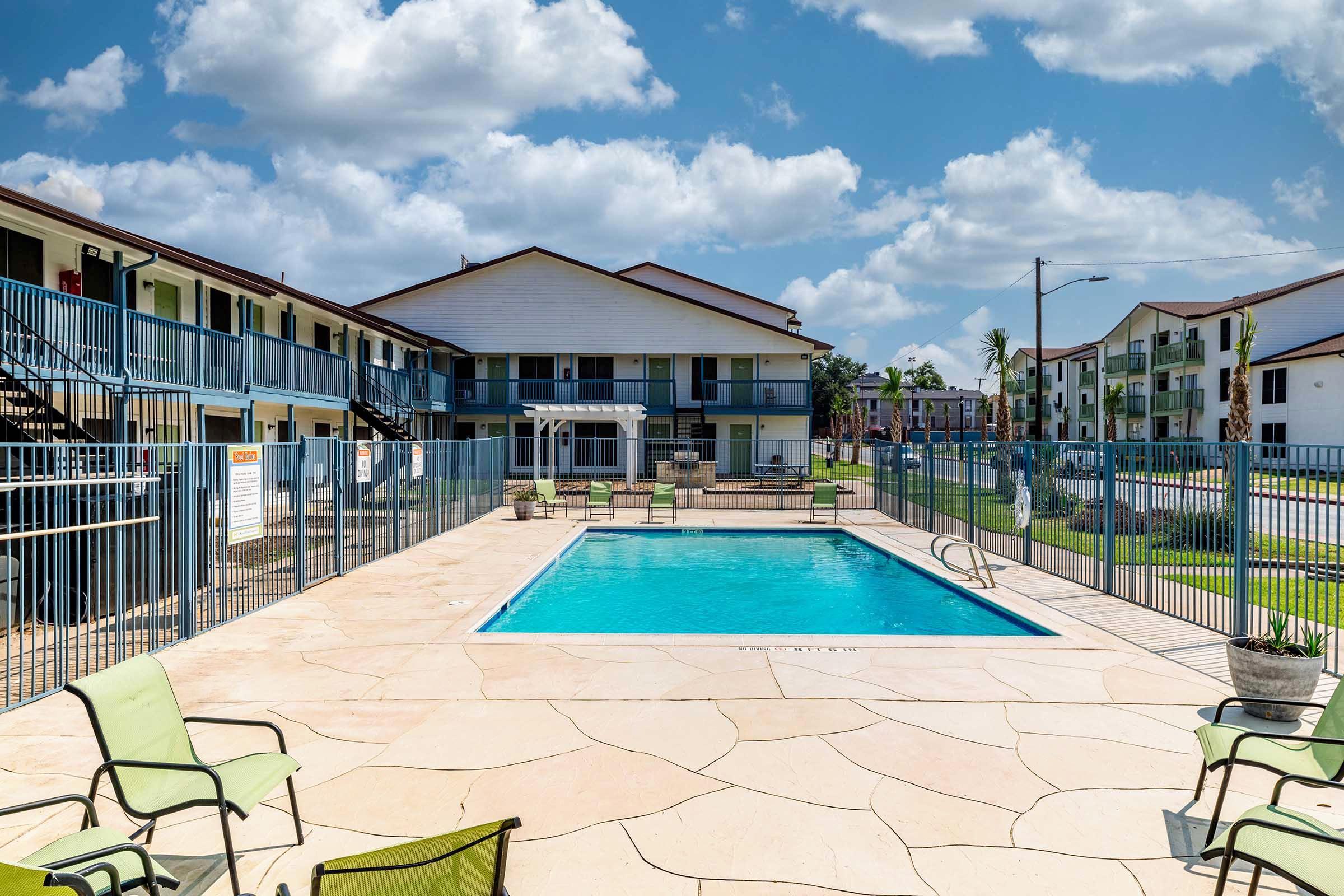 A sunny view of an outdoor swimming pool surrounded by a gated patio. Green lounge chairs are positioned nearby, and two-story apartment buildings are visible in the background against a partly cloudy sky.