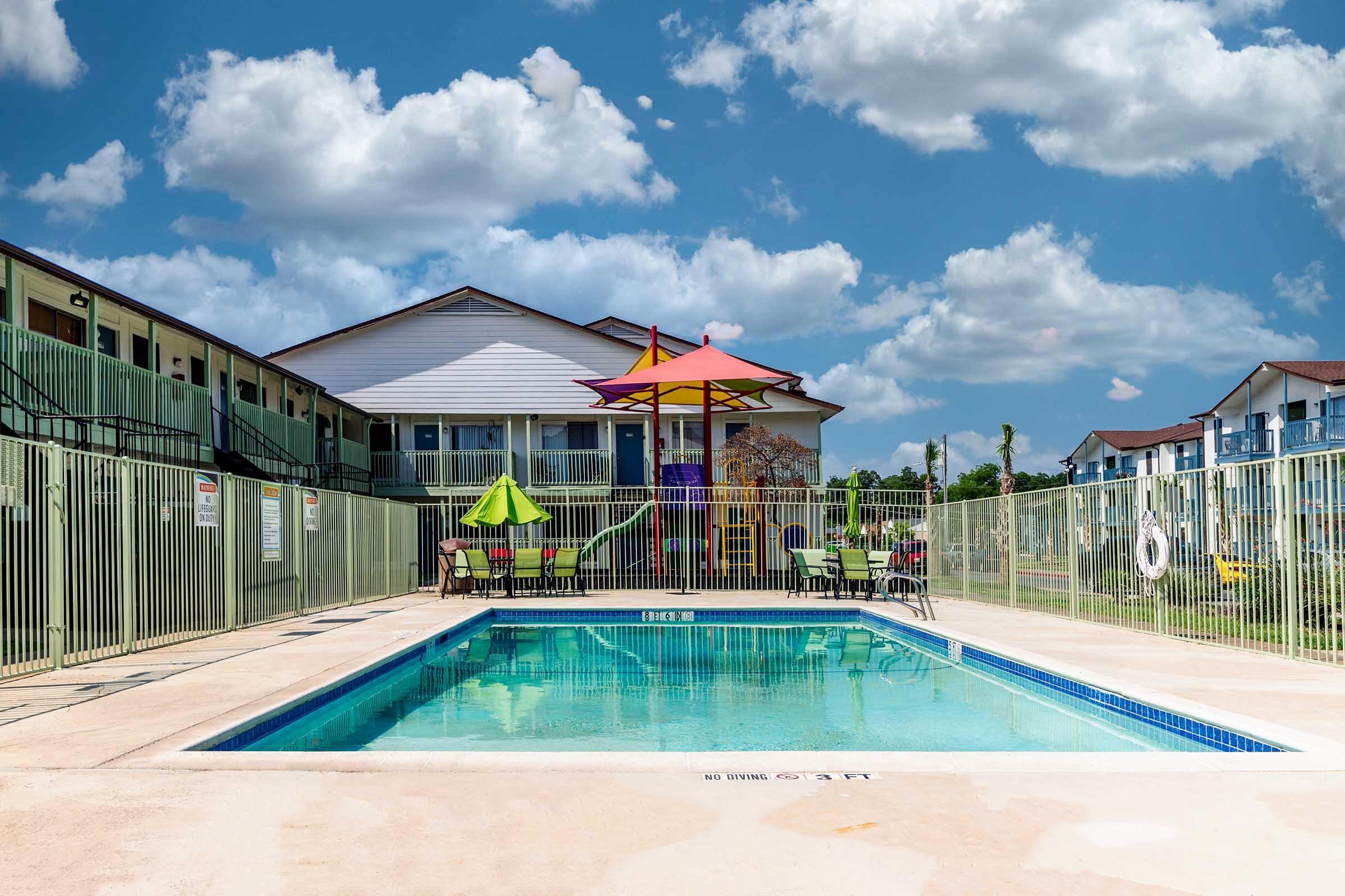 A swimming pool surrounded by a fenced area, featuring colorful umbrellas and lounge chairs. In the background, there's a two-story building with multiple units, and the sky is partly cloudy, creating a bright and inviting atmosphere.