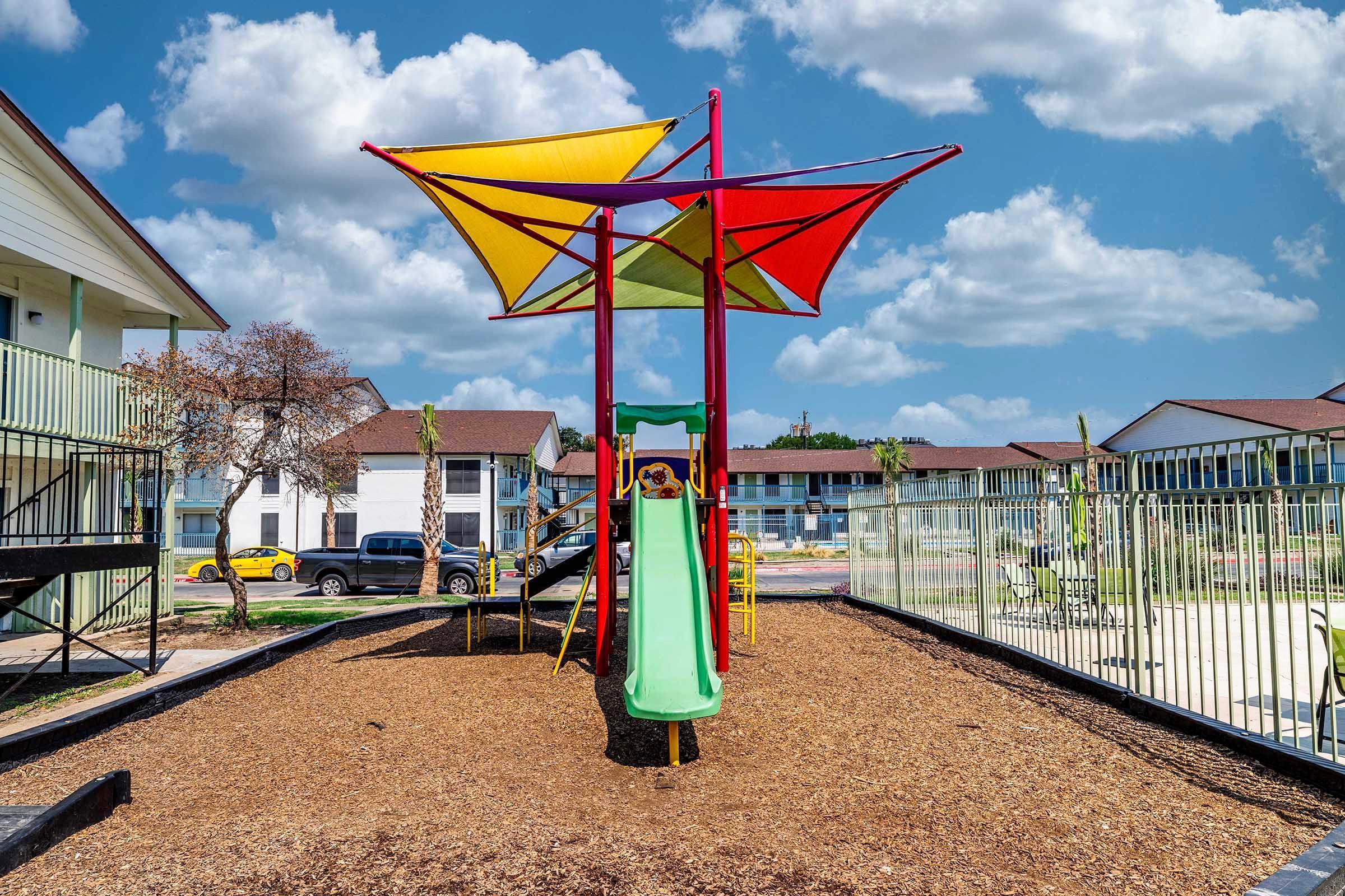A colorful playground featuring a slide and a large shaded canopy, surrounded by wood chips. The backdrop includes a few residential buildings under a blue sky with scattered clouds. The area is well-maintained, suggesting a vibrant community space for children to play.