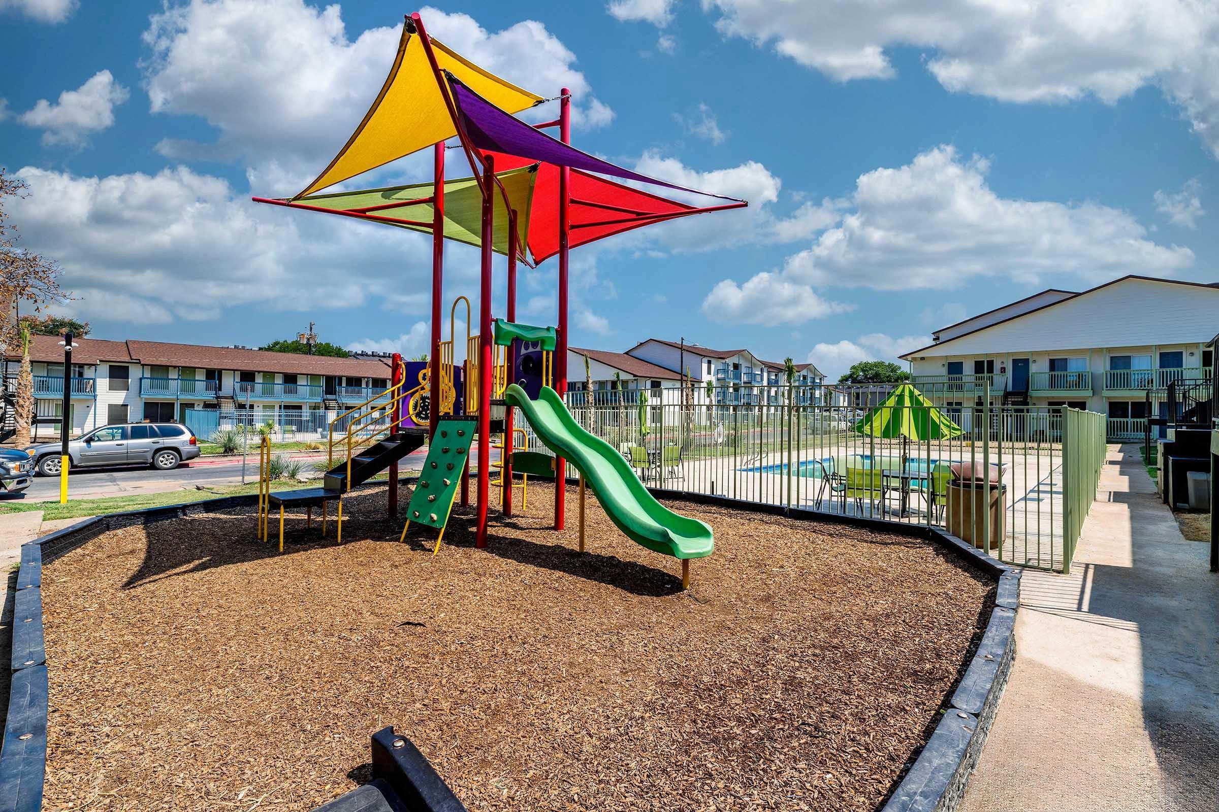 Playground featuring a colorful climbing structure with slides, surrounded by a wood chip area. In the background, there are two-story apartment buildings and a swimming pool area, under a blue sky with fluffy white clouds.