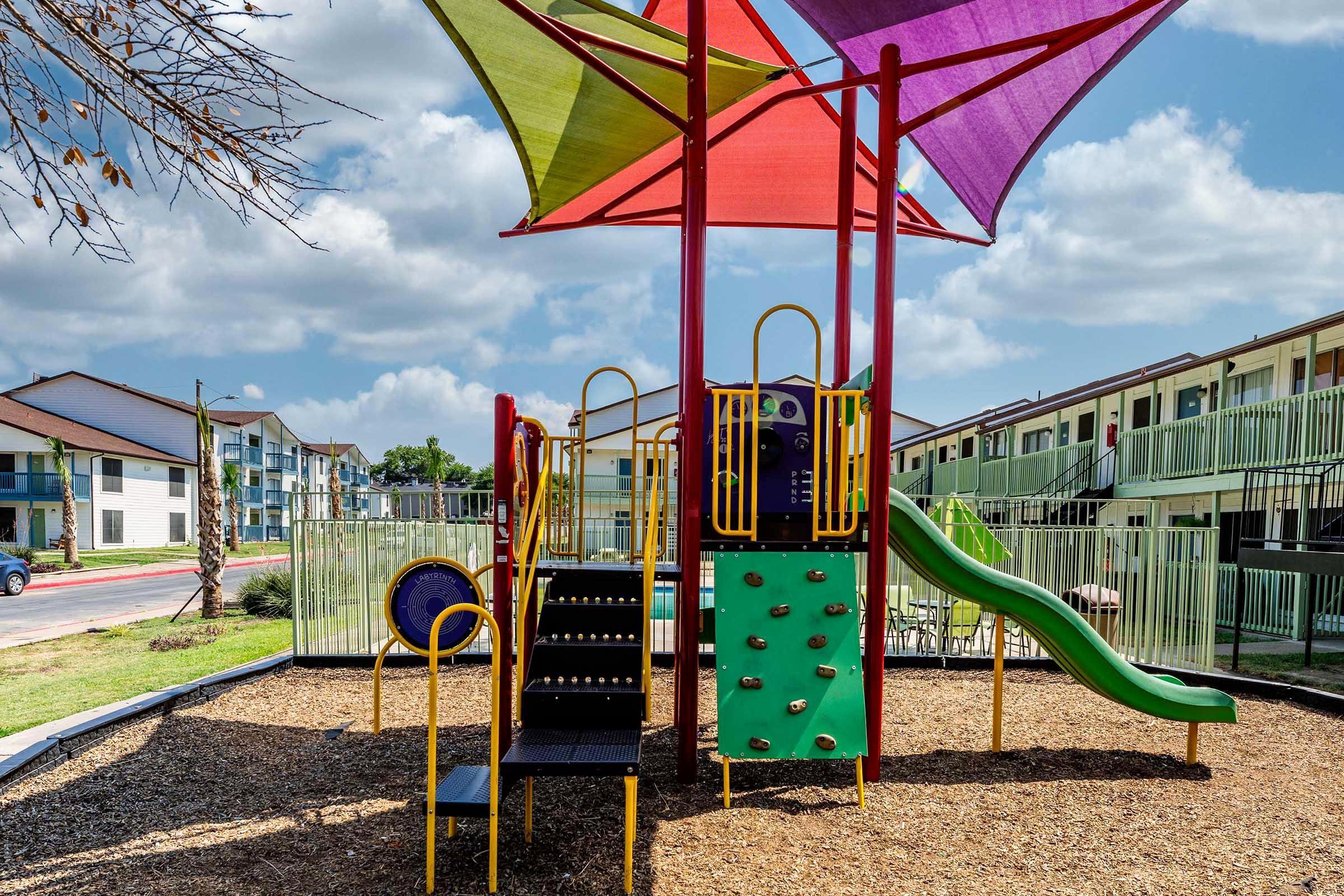 Colorful playground equipment with a slide, climbing wall, and shaded areas under large canopies, situated on a bed of mulch. In the background, multi-story residential buildings with balconies are visible under a partly cloudy sky.