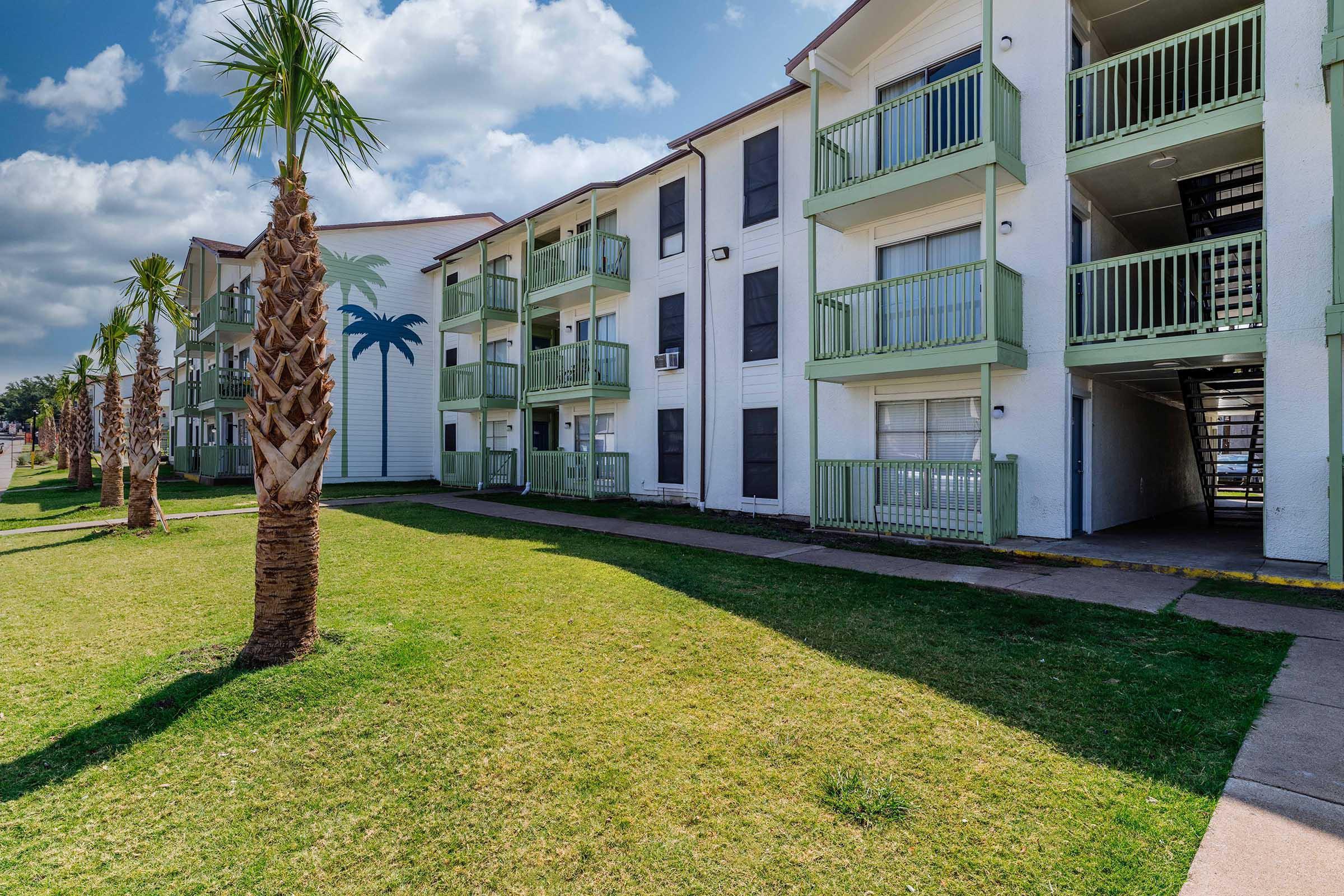 A residential apartment complex featuring multiple white buildings with green balconies, palm trees, and well-maintained landscaping. The scene is bright and sunny, capturing a peaceful outdoor environment.