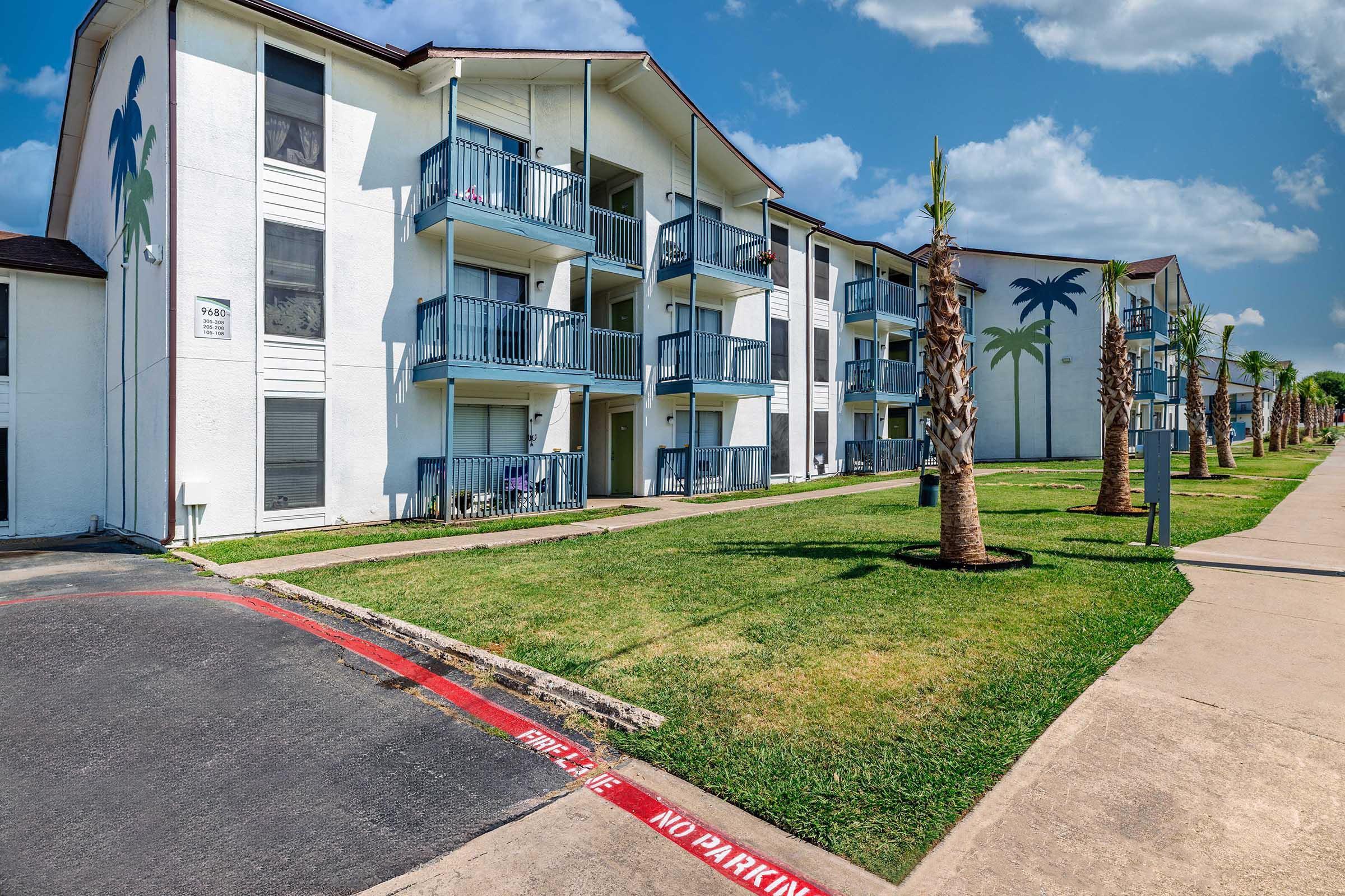A view of a multi-story apartment building with balconies and palm trees in a landscaped yard. The building features white walls and light blue railings, with a clear blue sky above. A "No Parking" sign is visible on the curbside. The scene conveys a modern, inviting residential atmosphere.