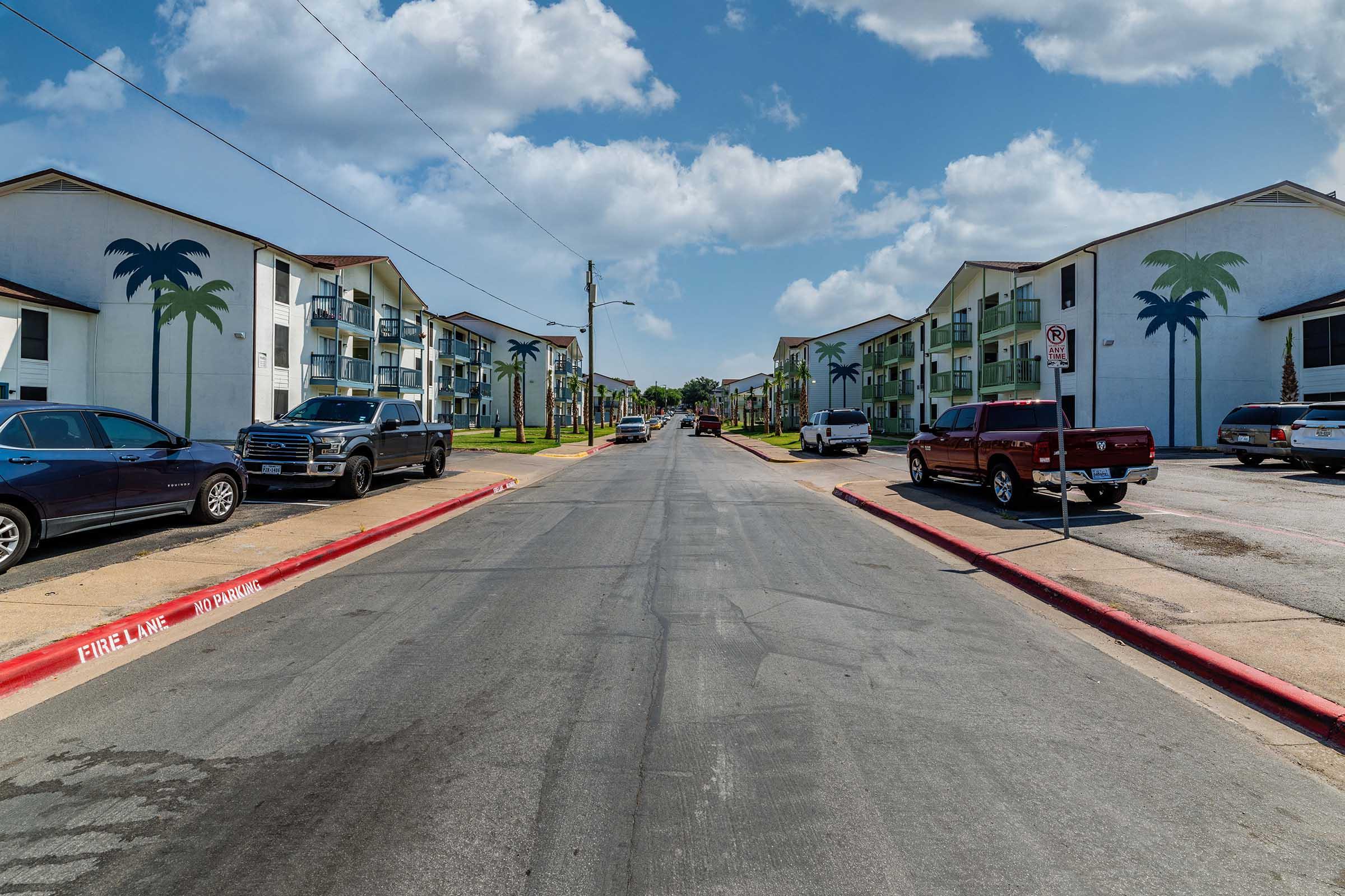 A residential street lined with parked cars on both sides, flanked by two-story apartment buildings adorned with palm tree murals. The sky is partly cloudy, and power lines run overhead. The road is well-maintained, with a red curb indicating a no-parking zone on one side.
