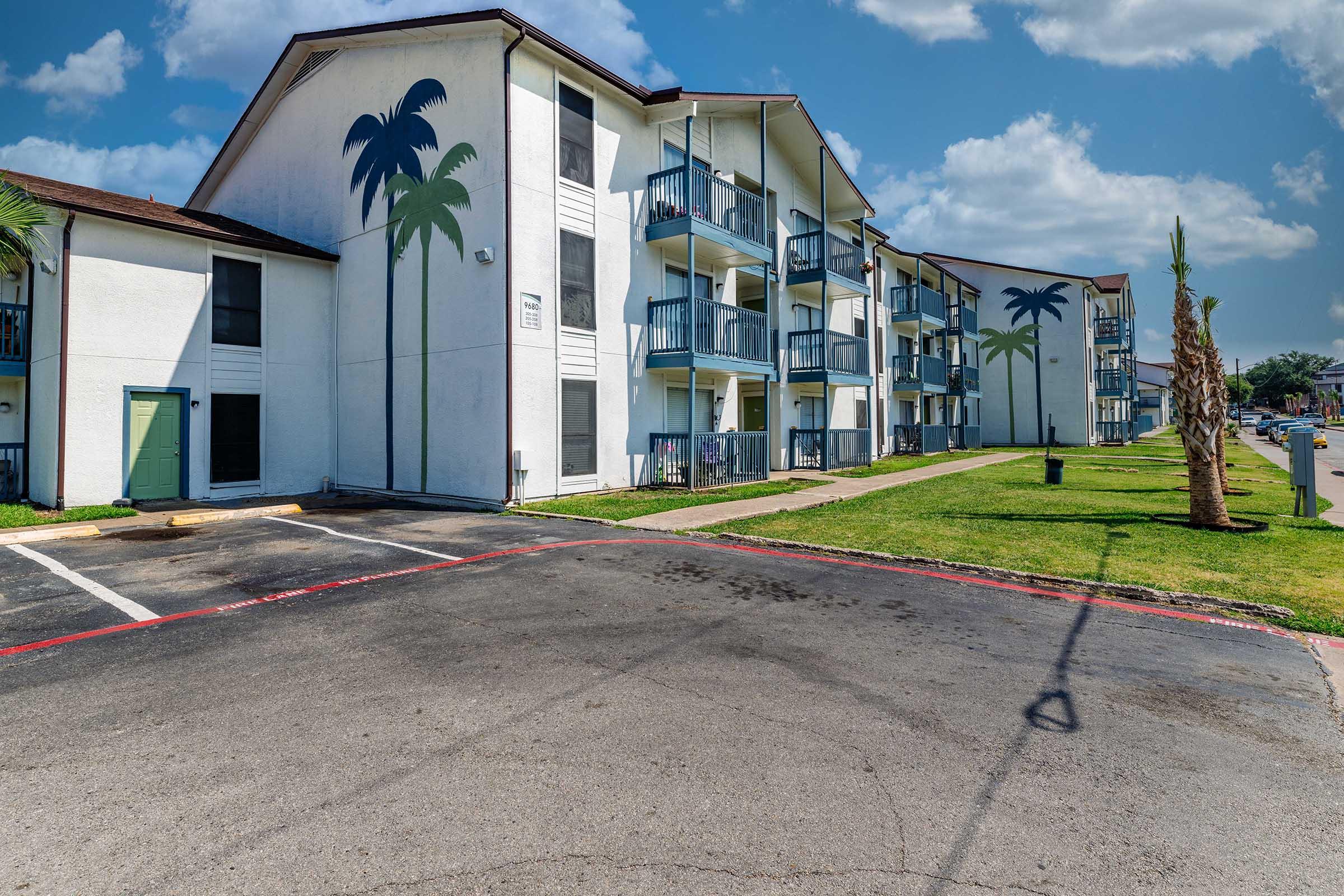 A view of a three-story apartment building with palm tree murals on the exterior. The building is surrounded by green grass and has a parking area in front. The sky is partly cloudy, creating a sunny atmosphere.