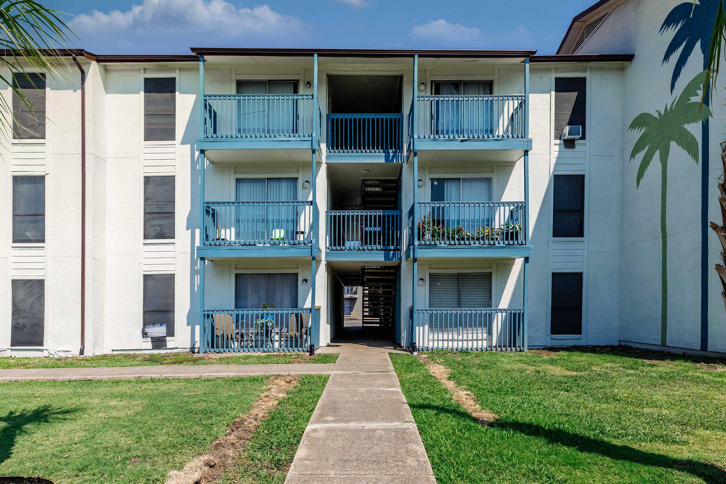 A three-story residential building with blue balconies. The front features a pathway leading to the entrance, surrounded by neatly manicured grass. Some balconies display potted plants, and there are palm trees on the right side, contributing to a tropical aesthetic. The sky is clear with a few clouds.