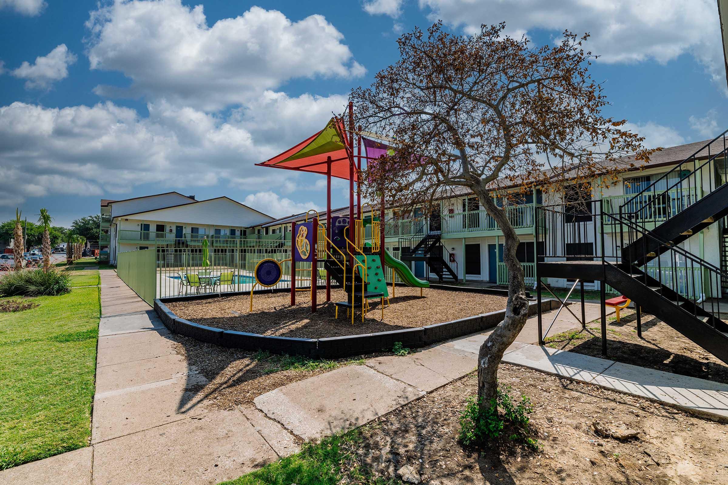 A colorful playground featuring slides and swings, situated in a grassy area surrounded by two-story apartment buildings. The sky is partly cloudy, and a small tree is visible nearby, enhancing the outdoor setting. The scene captures a cheerful environment for children.
