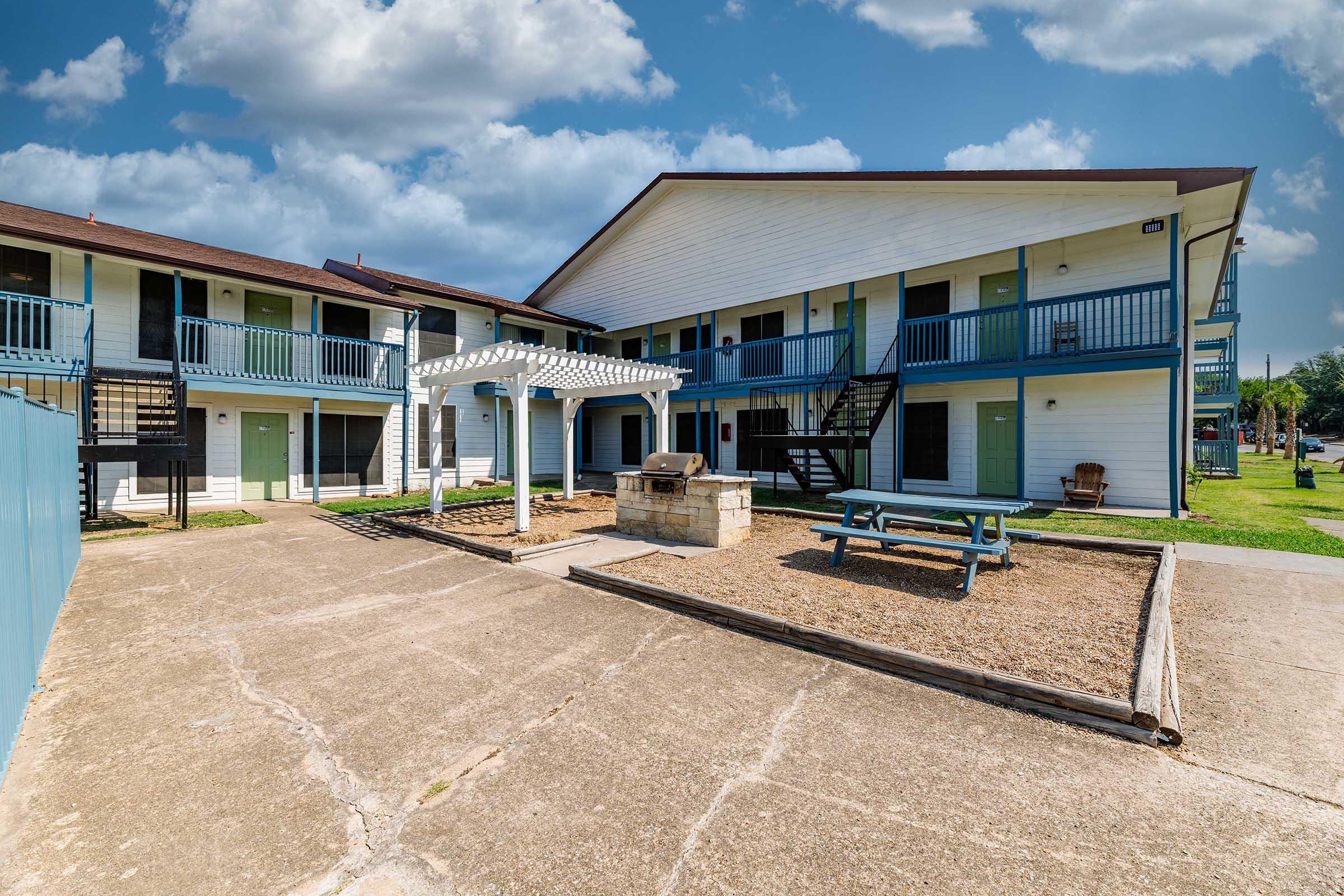 A two-story apartment building with balconies and a central courtyard featuring a picnic area, a stone grill, and landscaping. The sky is partly cloudy, and a grassy area surrounds the building, providing a pleasant outdoor space for residents.
