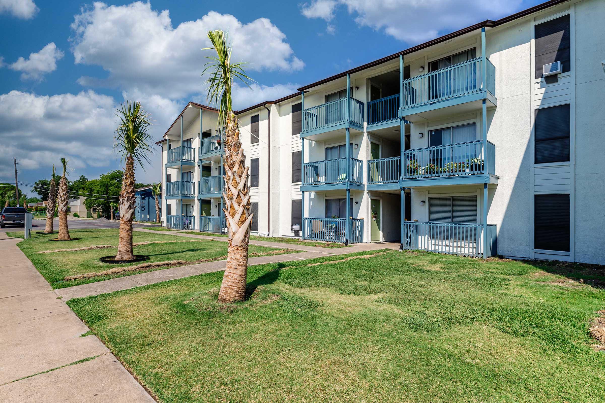 A multi-unit apartment building with two floors featuring balconies. Lush green grass lines the walkway, and palm trees are planted in front of the building. The sky is partly cloudy, creating a bright and inviting atmosphere.