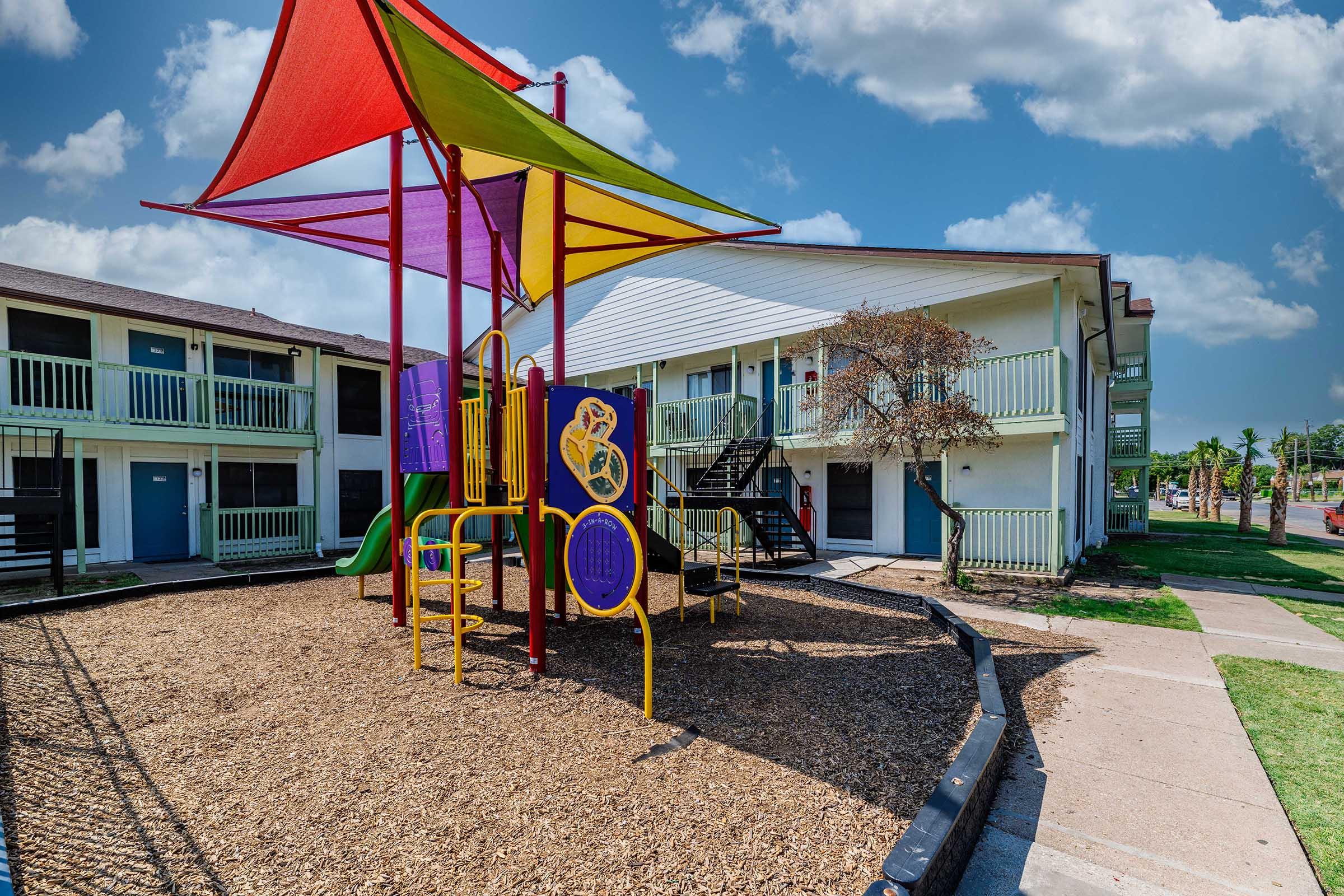 Colorful playground equipment with a large shaded structure stands on a bed of mulch, surrounded by grass. Nearby, two-story residential buildings with balconies are visible. The sky is partly cloudy, creating a bright and inviting atmosphere.