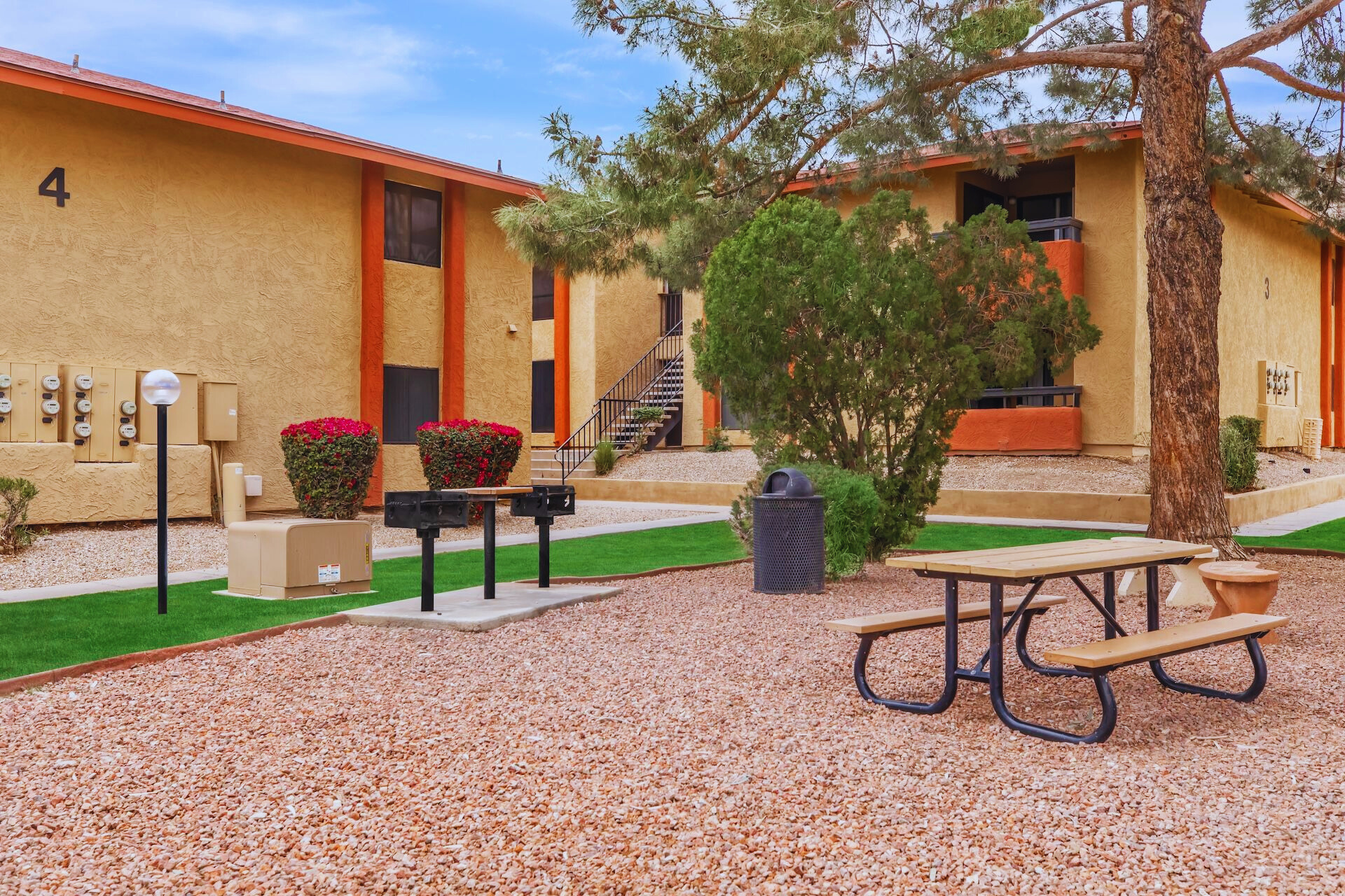 A landscaped courtyard featuring two apartment buildings with orange and beige exteriors. In the foreground, there is a picnic table and two charcoal grills on a pebbled area, surrounded by shrubs and trees. A trash bin is also visible, along with utility boxes mounted on the building.