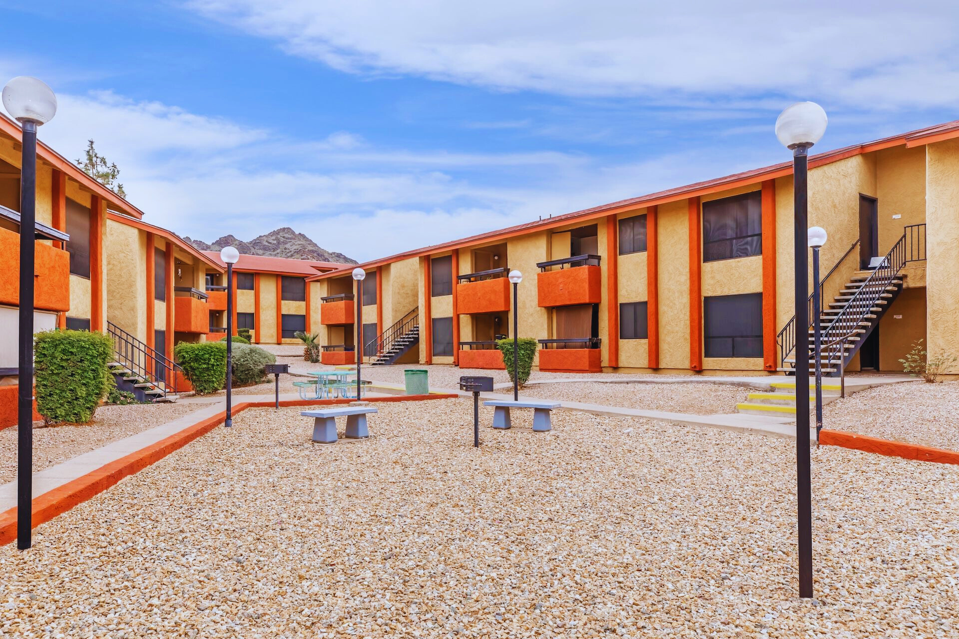 An outdoor view of a multi-story apartment complex with orange and yellow buildings. The courtyard features a sandy area with benches, walking paths, and lamp posts. In the background, there are mountains under a blue sky with clouds. The scene conveys a quiet and pleasant residential environment.