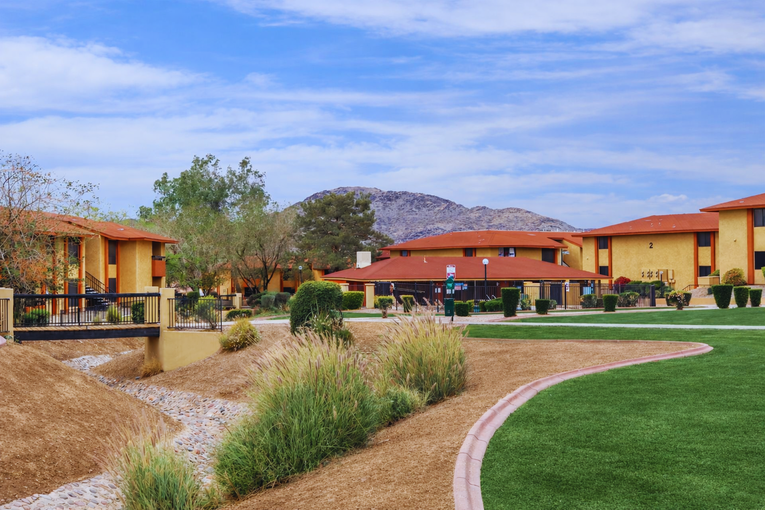 A landscaped residential area featuring low-rise buildings with orange roofs, green lawns, and well-maintained pathways. A small dry creek runs through the scene, surrounded by shrubs and trees, with a backdrop of rolling hills under a blue sky with scattered clouds.