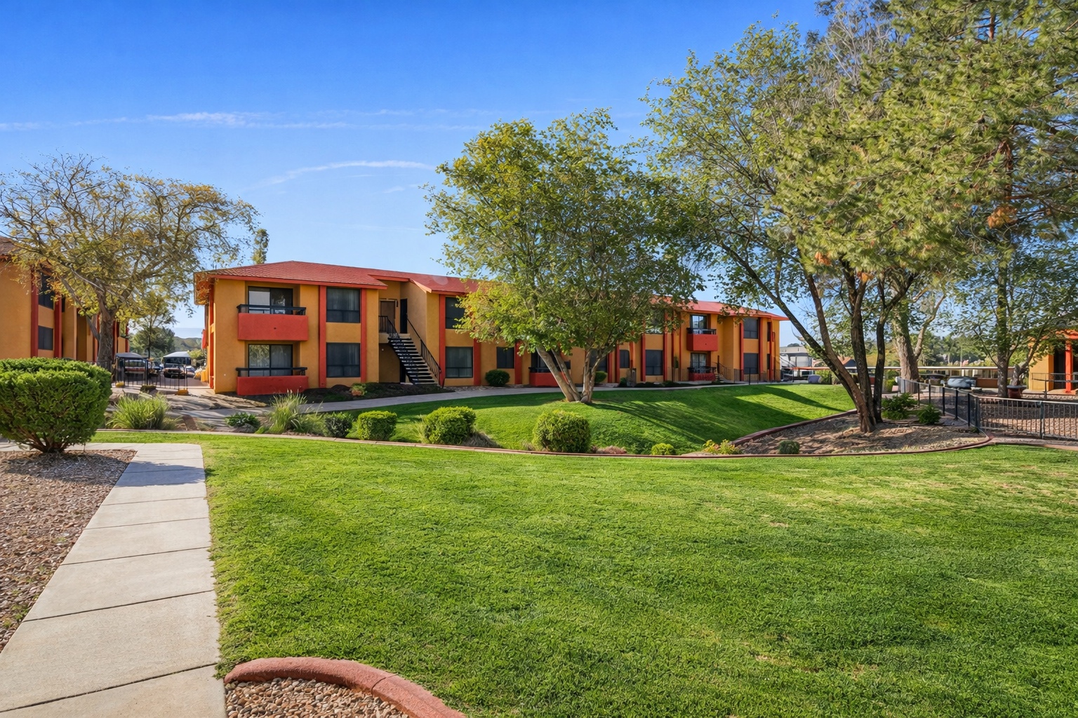 A well-maintained residential complex featuring orange and yellow buildings with balconies, surrounded by lush green lawns and trees. A paved walkway leads through the grassy area, and parking spaces are visible nearby, under a clear blue sky.