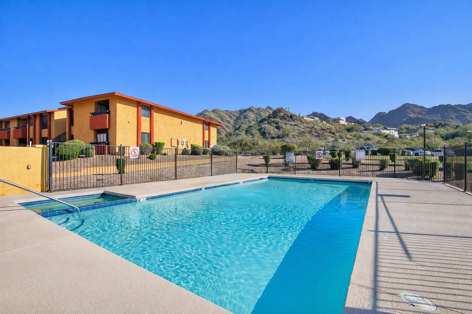 A clear swimming pool surrounded by a fence, with a vibrant blue sky above. In the background, there are desert mountains and a two-story building with a warm-colored facade. The pool area features lounge chairs and well-maintained landscaping, creating a serene atmosphere.