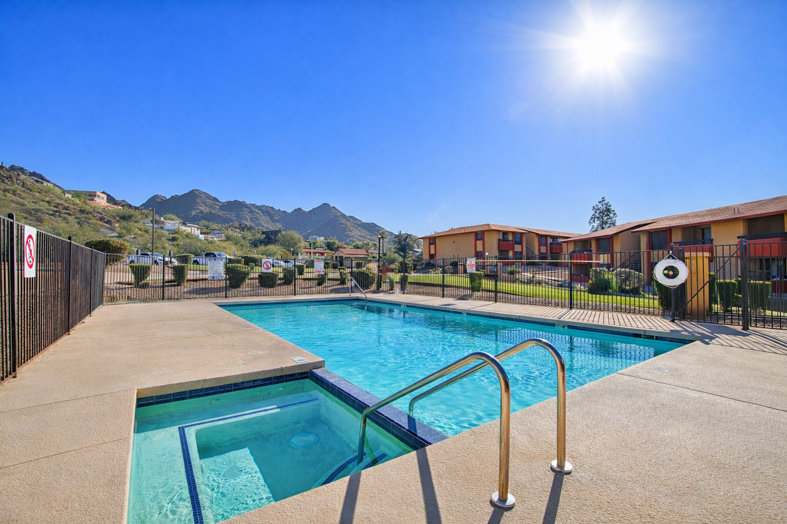 A clear blue swimming pool with a hot tub, surrounded by a metal fence. In the background, there are mountains and buildings. The sun is shining brightly in a clear blue sky. The pool deck is well-maintained, with a ladder leading into the pool.