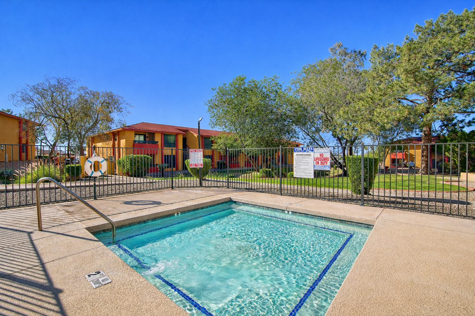 A clear hot tub sits beside a swimming pool, surrounded by a black iron fence. In the background, colorful apartment buildings are visible, along with trees and landscaping. A blue sky complements the scene, adding to the inviting atmosphere of the outdoor space.