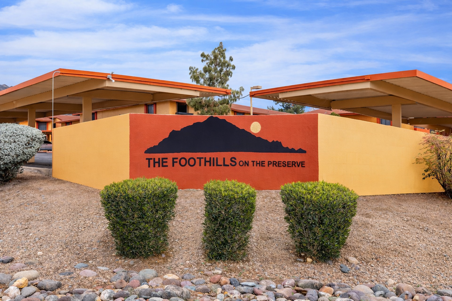 A colorful sign reading "THE FOOTHILLS ON THE PRESERVE" with a mountain silhouette and sun icon. The sign is surrounded by well-maintained shrubs and gravel, set against a backdrop of modern-style buildings under a clear blue sky.