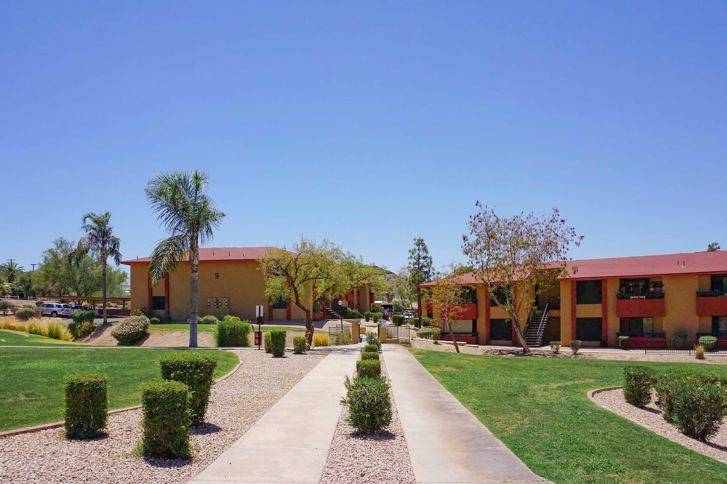 A sunny outdoor view of a well-maintained residential complex with two-story buildings. The pathway is lined with neatly trimmed hedges and decorative gravel, surrounded by green lawns and a few trees. Clear blue sky overhead completes the serene atmosphere.