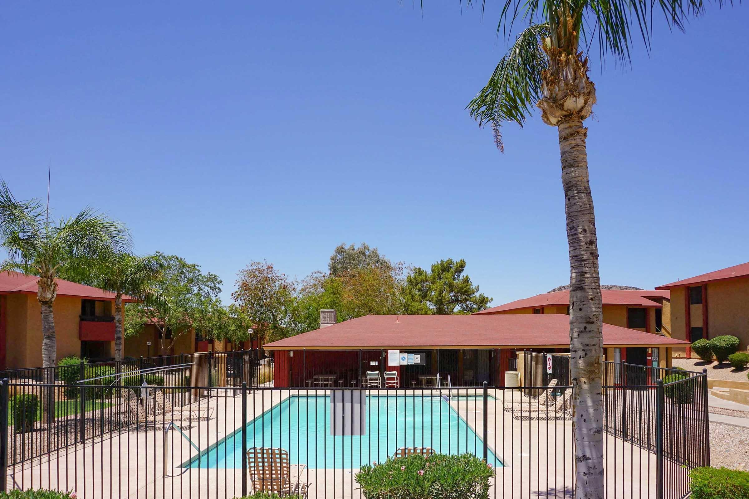 A clear blue sky overhead with a swimming pool surrounded by a gated area. Palm trees and shrubs are visible, and two single-story buildings with red roofs are situated on either side of the pool area. Sun loungers are arranged around the pool, creating a relaxing outdoor environment.