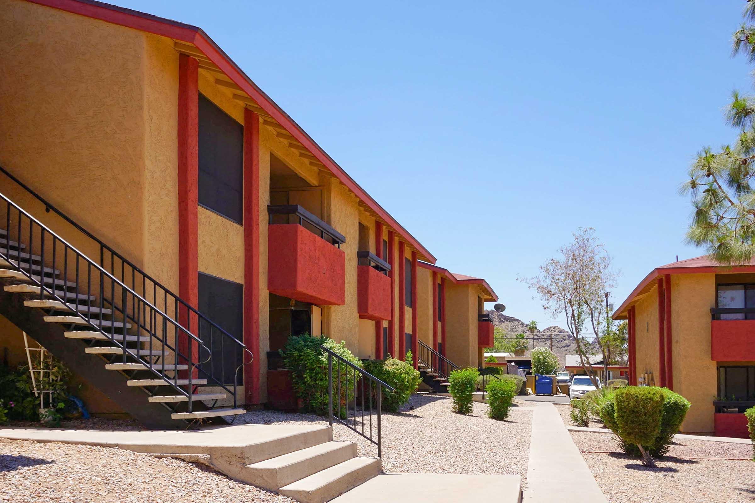 A view of two-story apartment buildings with red accents. A concrete pathway leads through landscaped areas with small bushes. Metal staircases are visible on one side, and there are trees in the vicinity against a clear blue sky. The scene suggests a warm, sunny day.