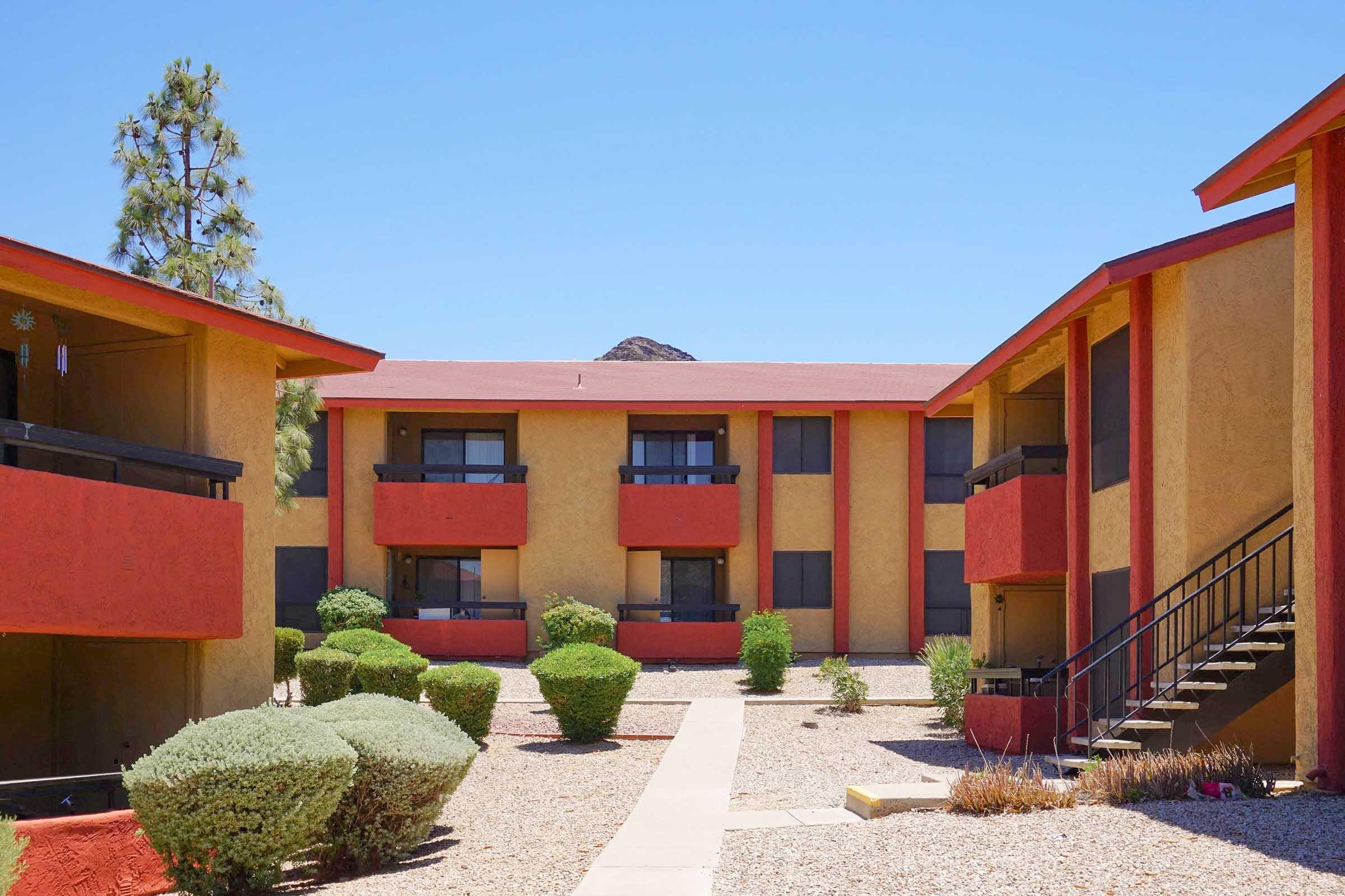 View of a multi-story apartment community with a vibrant color scheme, featuring red and yellow exteriors. The courtyard is landscaped with bushes and gravel pathways, positioned under a clear blue sky, with a mountain visible in the background.