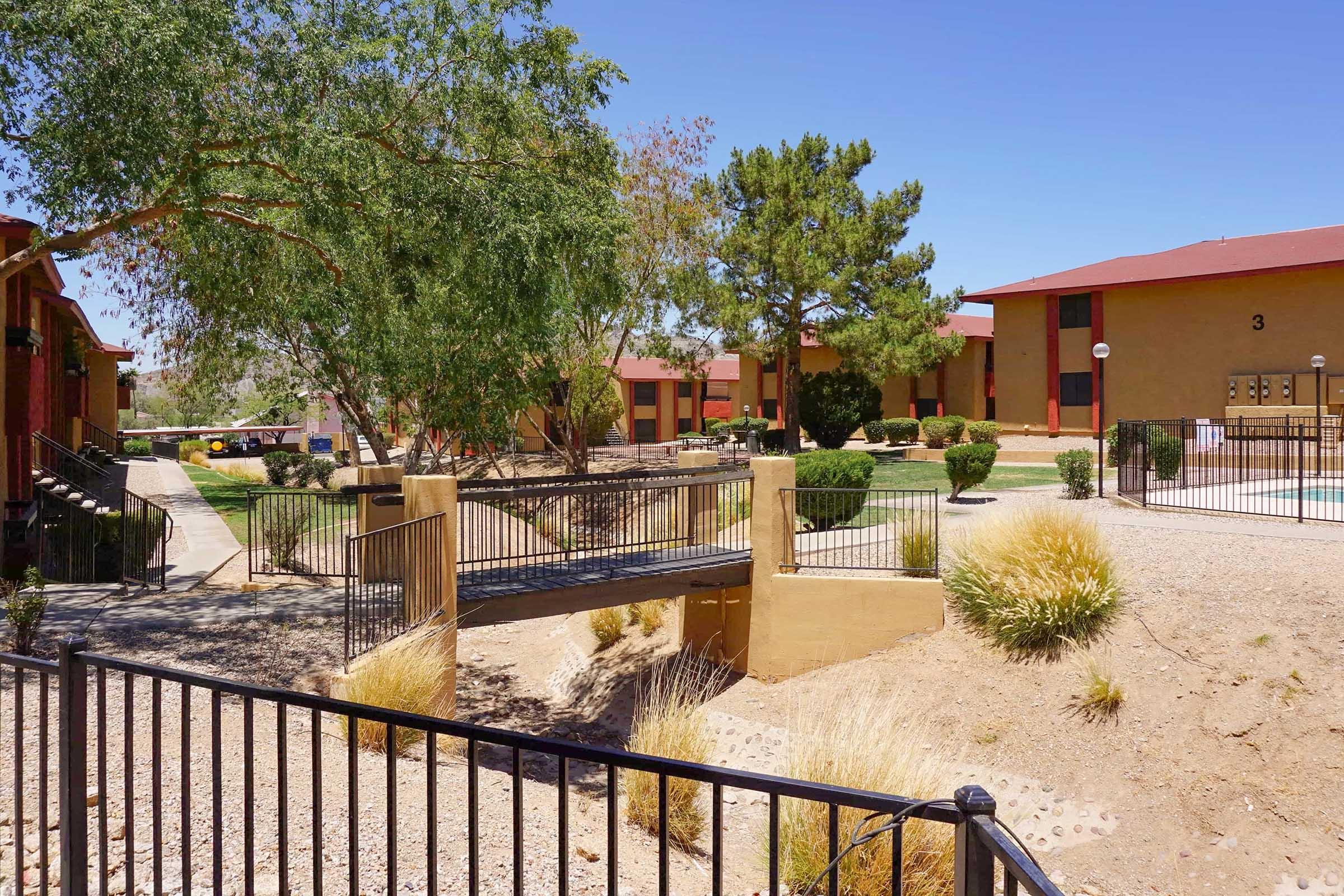 View of a residential complex featuring brown buildings with red roofs, a small footbridge over a dry stream, landscaped areas with shrubs and trees, and a clear blue sky in the background. The scene conveys a sunny, tranquil atmosphere in a well-maintained outdoor space.