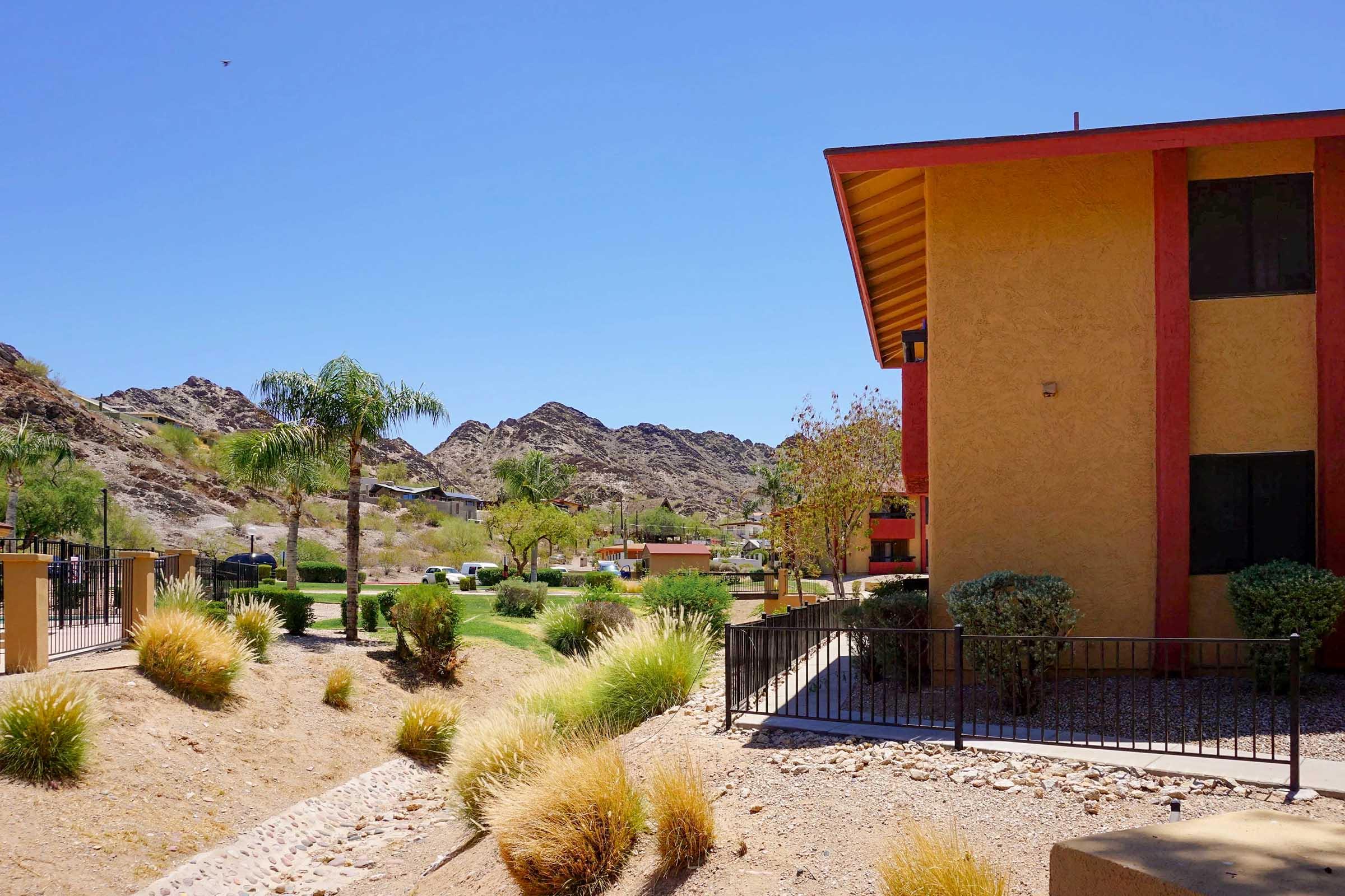 A bright, sunny landscape featuring a building with warm-colored exterior. In the foreground, desert plants and grasses line a pathway, while in the background, rugged mountains and trees create a scenic backdrop. The sky is clear blue, indicating a beautiful day.