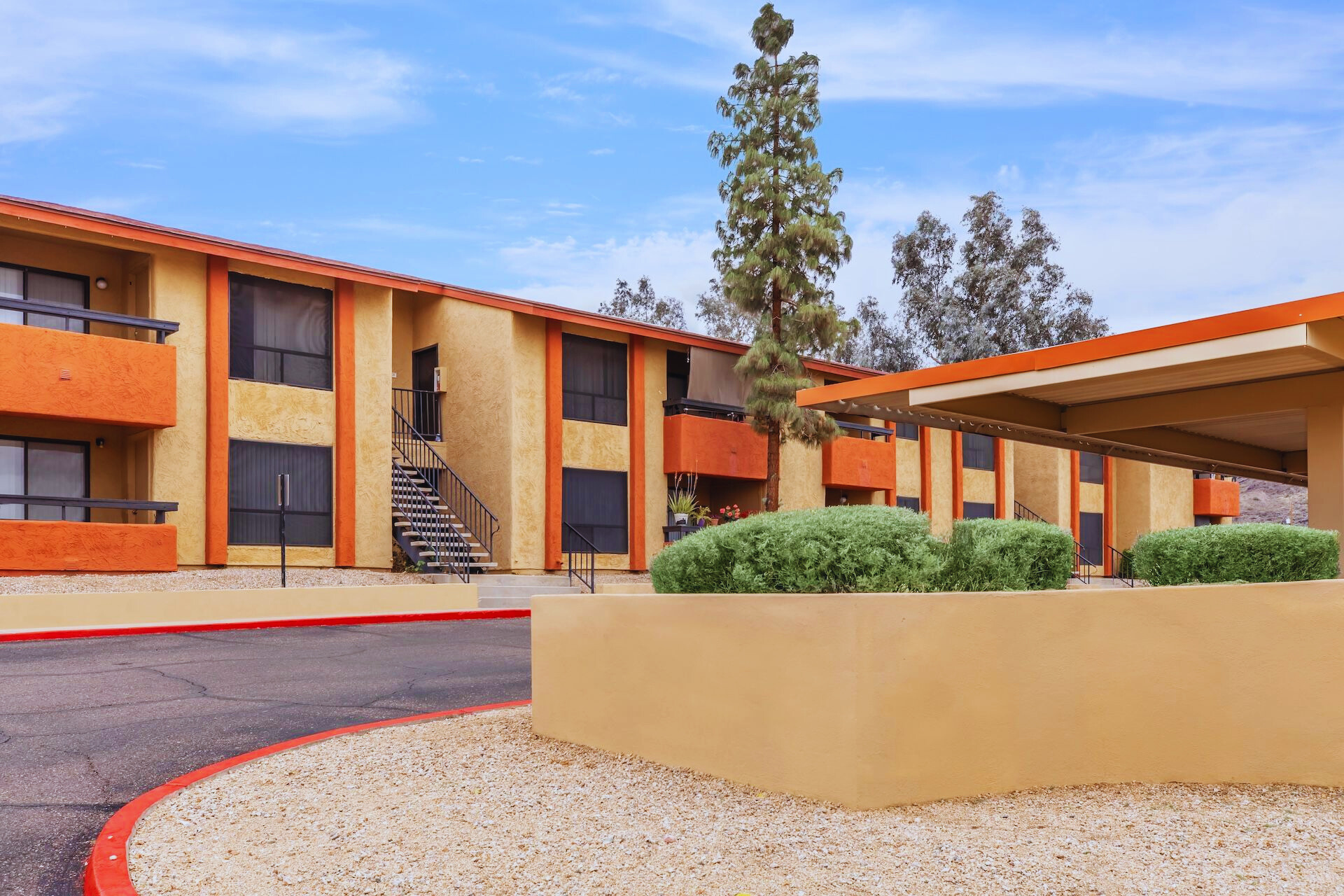 A modern low-rise apartment complex with orange and yellow exterior walls, featuring multiple units with balconies. There are landscaped bushes and a round driveway in the foreground, with a tree visible beside the building, against a blue sky with scattered clouds.