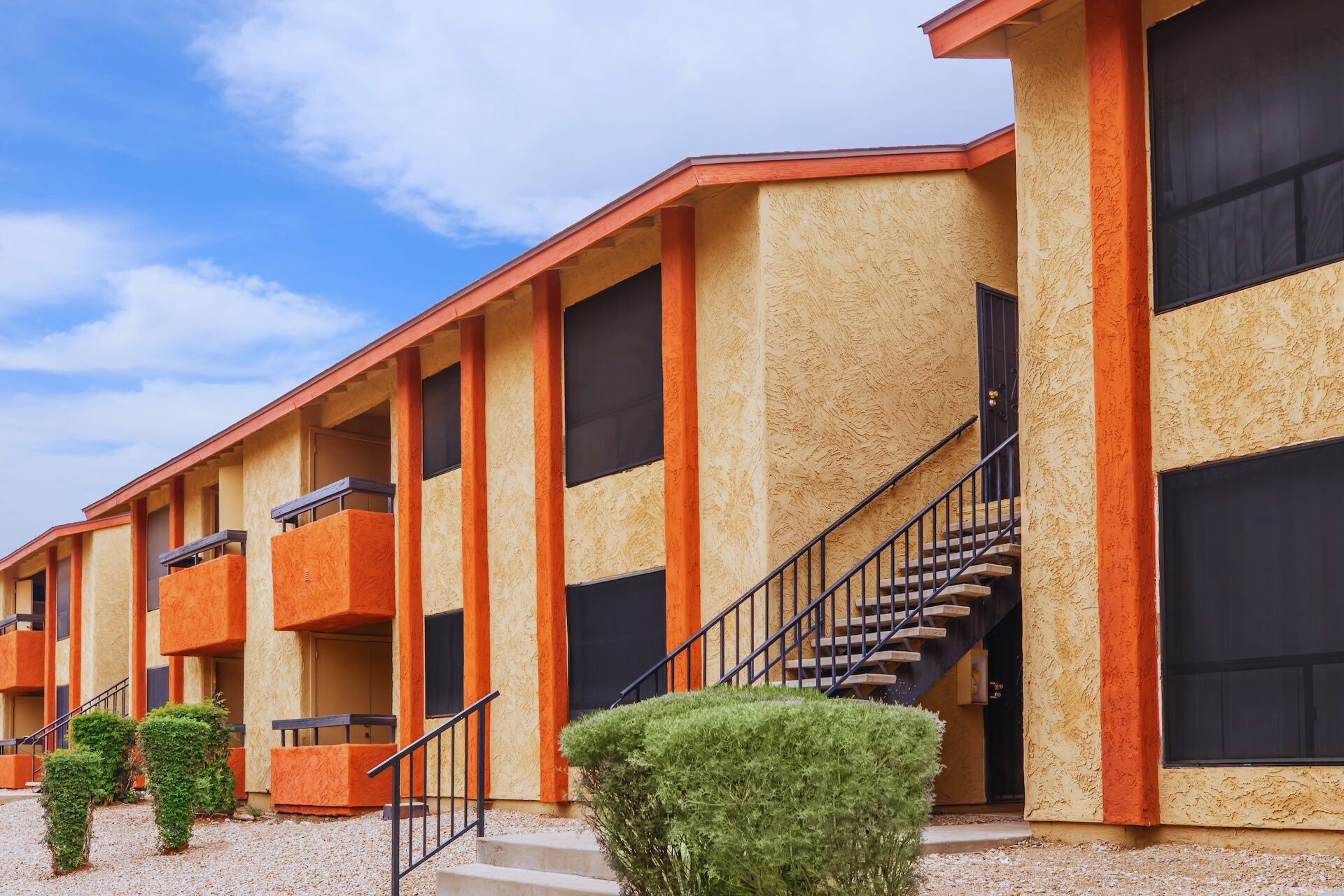 A view of an apartment building featuring orange and beige exterior walls. The building has multiple levels with balconies and staircases, surrounded by landscaped shrubs and gravel. The sky is partly cloudy, adding to the vibrant atmosphere of the scene.