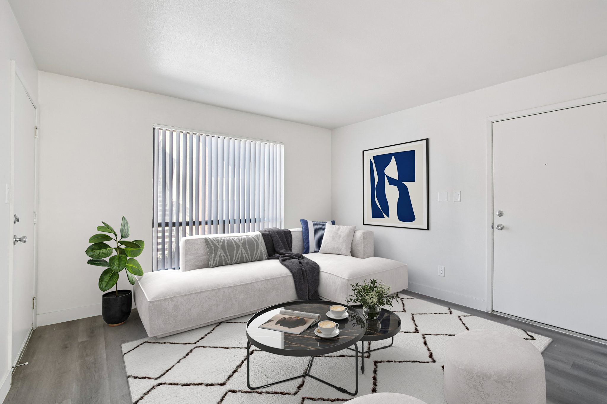A modern living room featuring a light-colored sectional sofa with decorative pillows, a round glass coffee table with tea cups, and a small plant. There is a large artwork on the wall in blue and white, a window with vertical blinds, and a minimalist décor style.