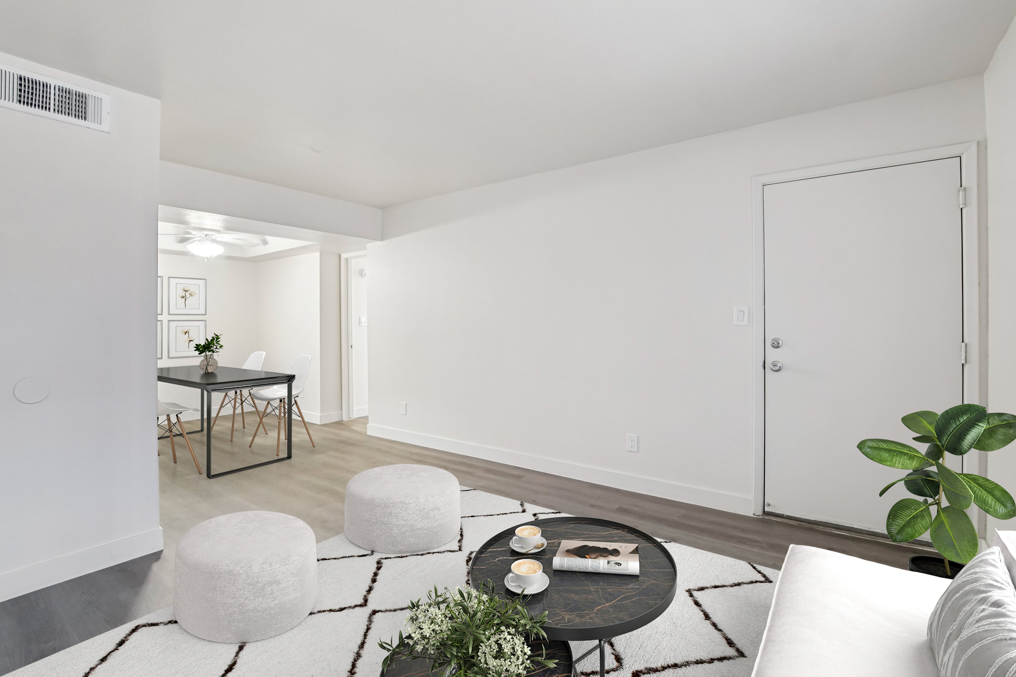 A modern, minimalist living space featuring a light-colored sofa with two round ottomans, a coffee table with a small plant and magazines, and a door leading to another room. A dining area with a table and chairs is visible in the background, and the walls are painted in neutral tones.