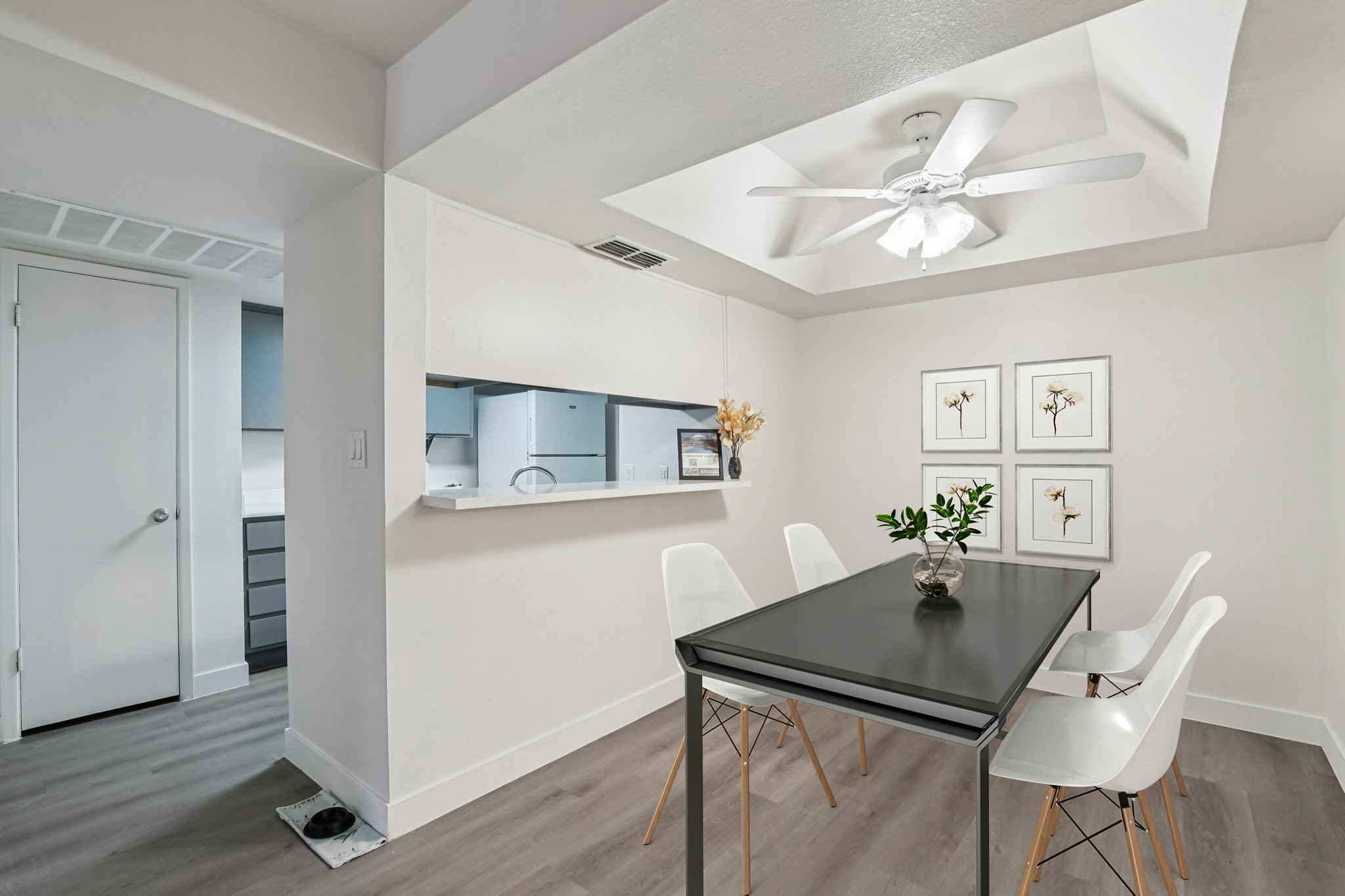 A modern dining area featuring a sleek black table surrounded by white chairs. A ceiling fan is installed above, and the room has light-colored walls. There are three framed flower prints on the wall, and an open kitchen area is visible in the background. The flooring is a light wooden style.