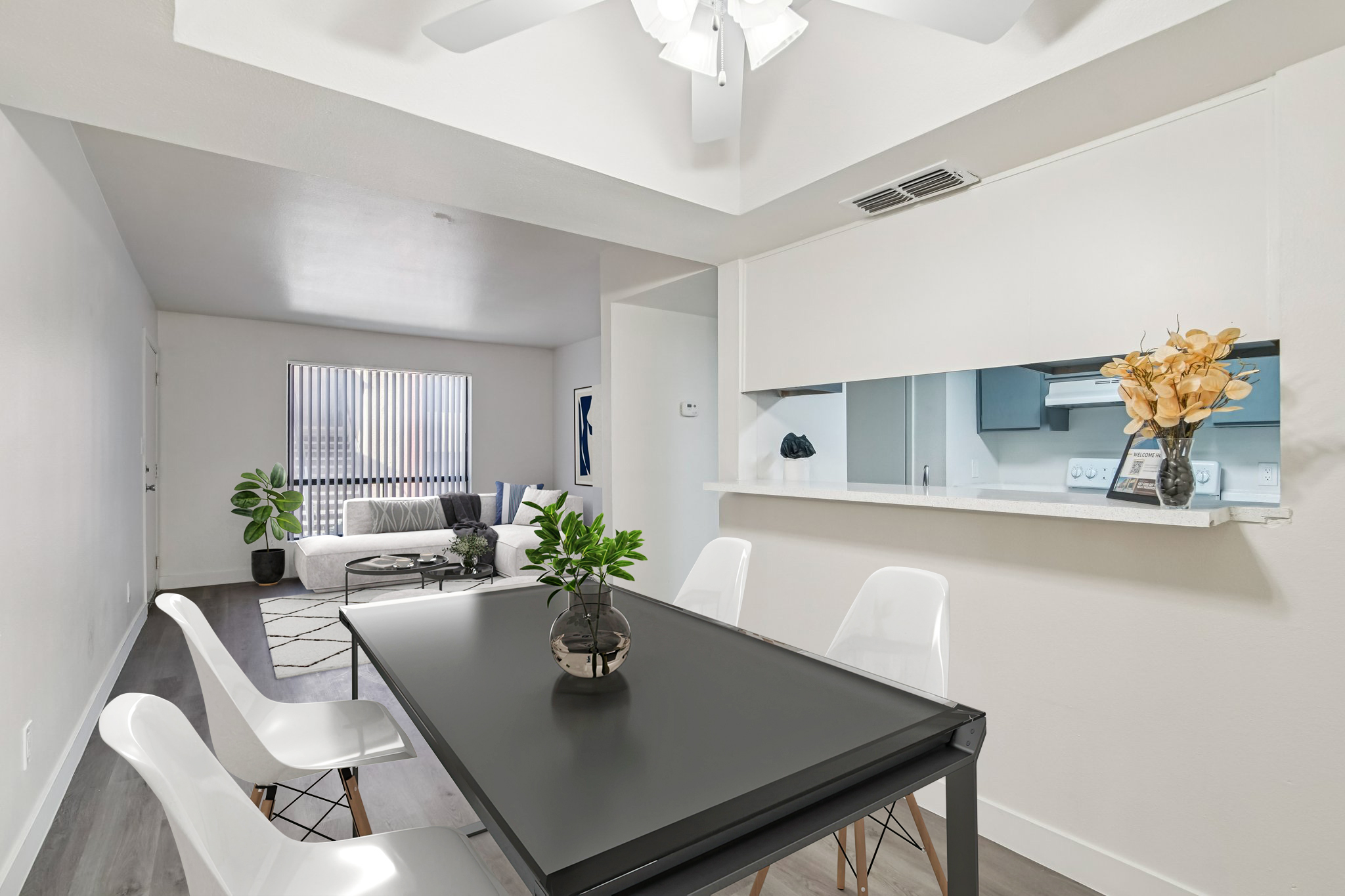 A modern dining area featuring a black table with white chairs, a decorative vase, and a small potted plant. In the background, a living room is visible with a light-colored sofa, large window with blinds, wall art, and a rug on the floor. The space is bright and airy with neutral tones.