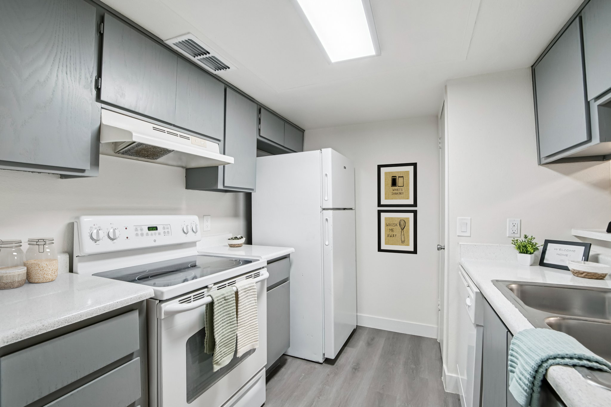 A modern kitchen featuring gray cabinetry, a white refrigerator, and an electric stove with a vent hood. The countertop is light and includes decorative items. Framed art is mounted on the wall, and the floor has a wood-like finish, creating a clean and inviting atmosphere.
