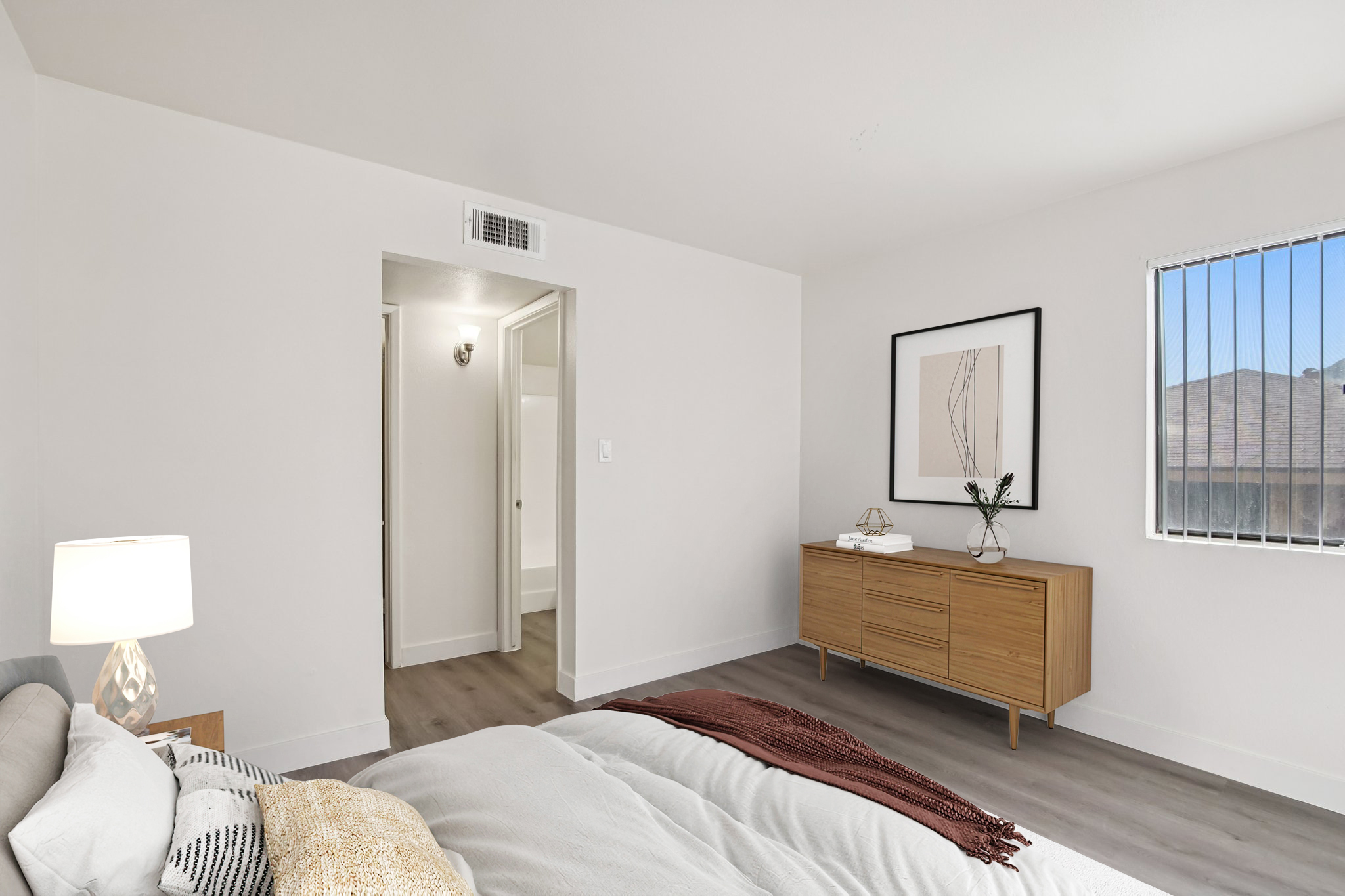 A modern, minimalist bedroom featuring a queen-sized bed with decorative pillows and a warm blanket. There's a nightstand with a lamp, a wooden dresser against the wall, and a framed artwork. A doorway leads to another room, and a window provides natural light. The walls are painted light and the floor is wood-like laminate.