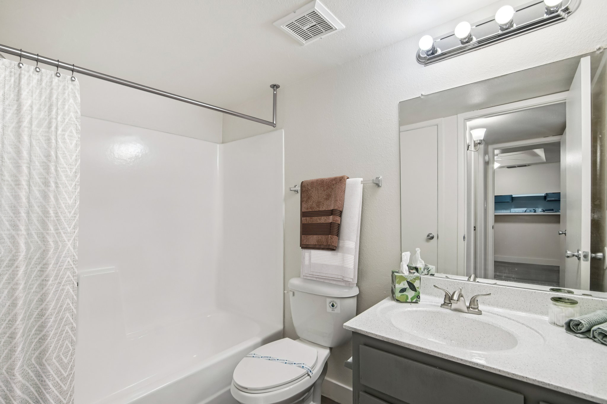 A modern bathroom featuring a bathtub with a shower, a toilet, and a vanity with a sink. The walls are painted in a neutral color, and the space is illuminated by overhead lights. Decorative towels hang on a towel rack, and there are small decorative items on the countertop.
