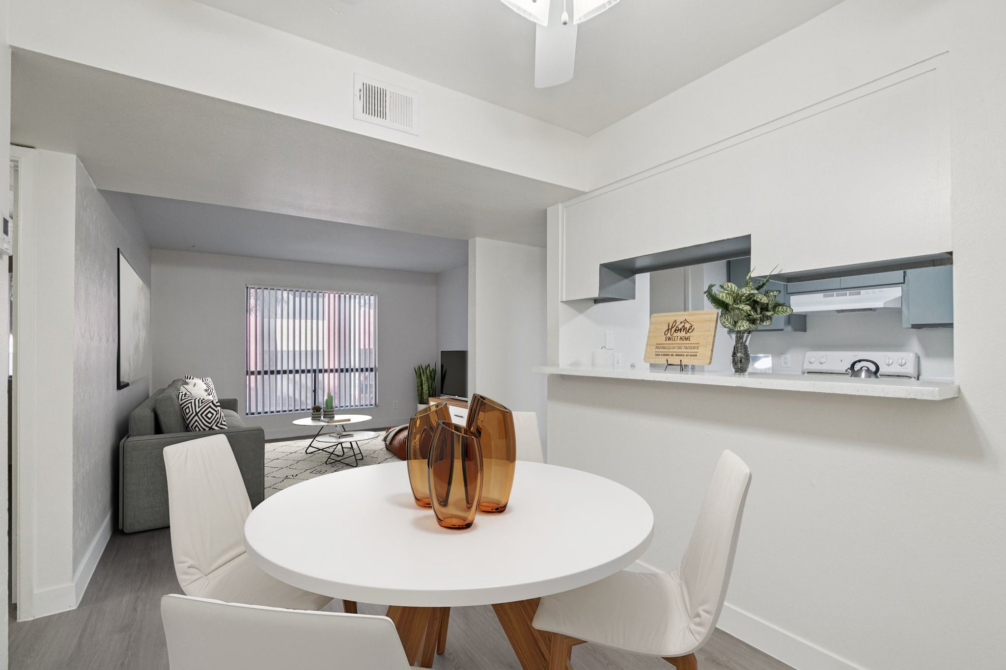 A modern, bright dining area featuring a circular white table with four white chairs. A decorative vase with various shapes sits in the center of the table. In the background, a cozy living room with a gray sofa and large window with vertical blinds is visible, adding to the contemporary aesthetic.
