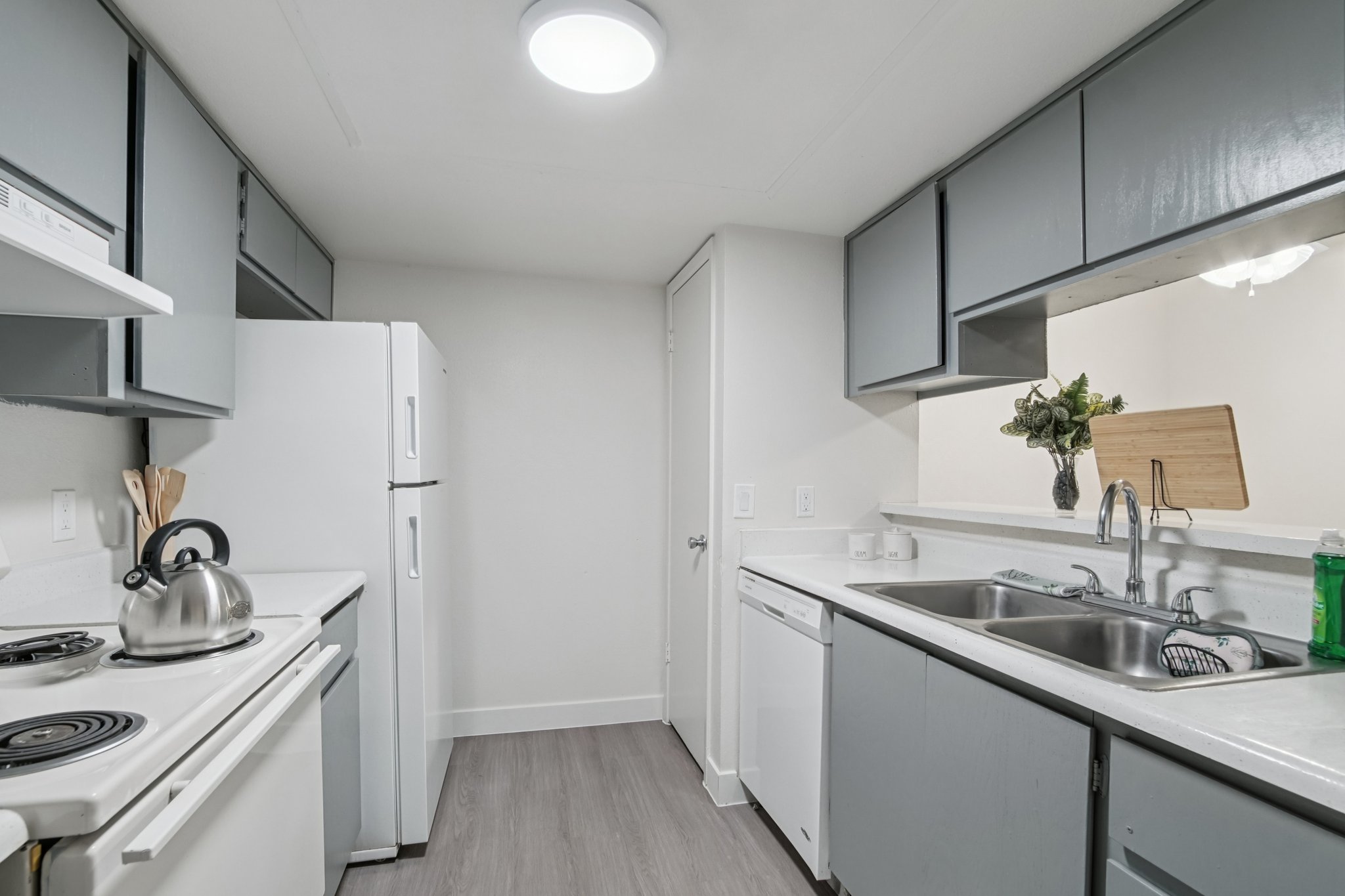 A modern kitchen featuring light gray cabinets, a white refrigerator and stove, stainless steel sink, and a dishwasher. The countertops are white with a simple design, and there's a small vase with greenery on the counter. The flooring is a light wood look, and the walls are painted a neutral color.