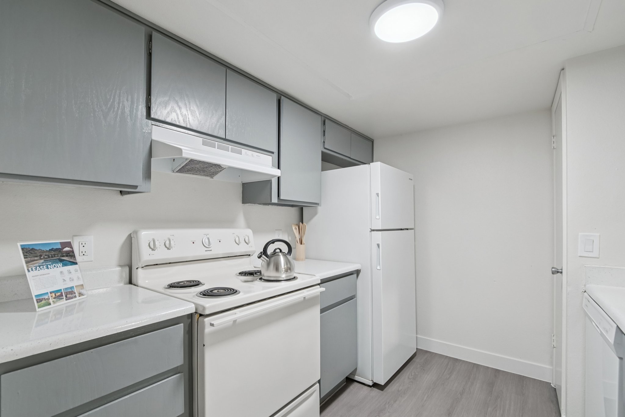 A modern kitchen with gray cabinetry, a white stove, a white refrigerator, and a stainless steel kettle on the stove. The countertop is light-colored, and there is a small display with a brochure on the left. The space is well-lit with a circular ceiling light and has a clean, minimalist design.