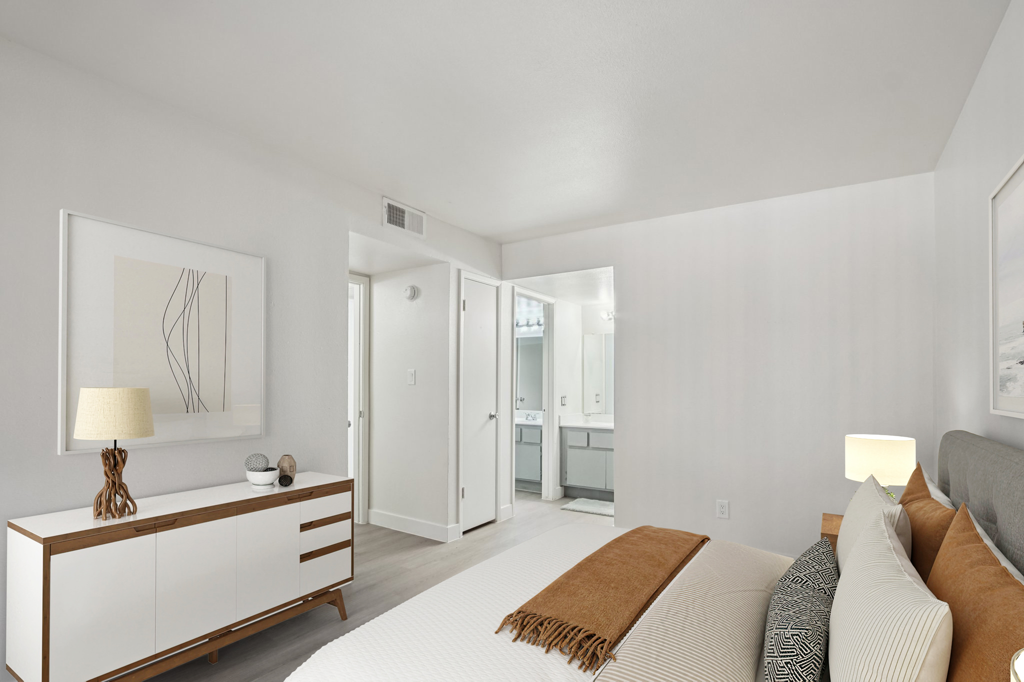A modern bedroom featuring a light color palette with a plush bed adorned with a tan throw. To the left, there is a stylish dresser with decorative items and a lamp. A doorway leads to a bathroom area, and the room is well-lit with natural light, creating a serene atmosphere.