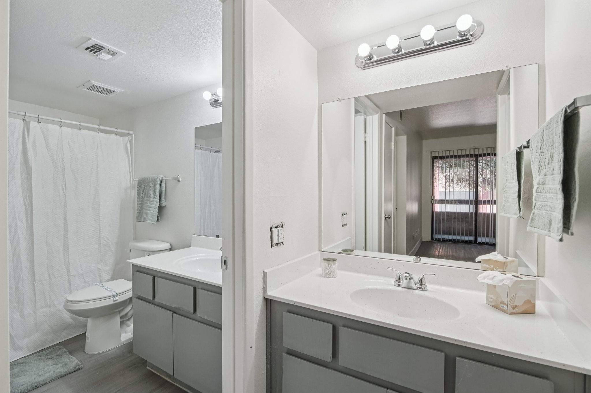 A clean, modern bathroom featuring a double vanity with gray cabinets, a large mirror, and a bright overhead light. The space includes a white shower curtain, a toilet, and a window with light filtering in. Soft-colored towels and a decorative item are placed on the counter.