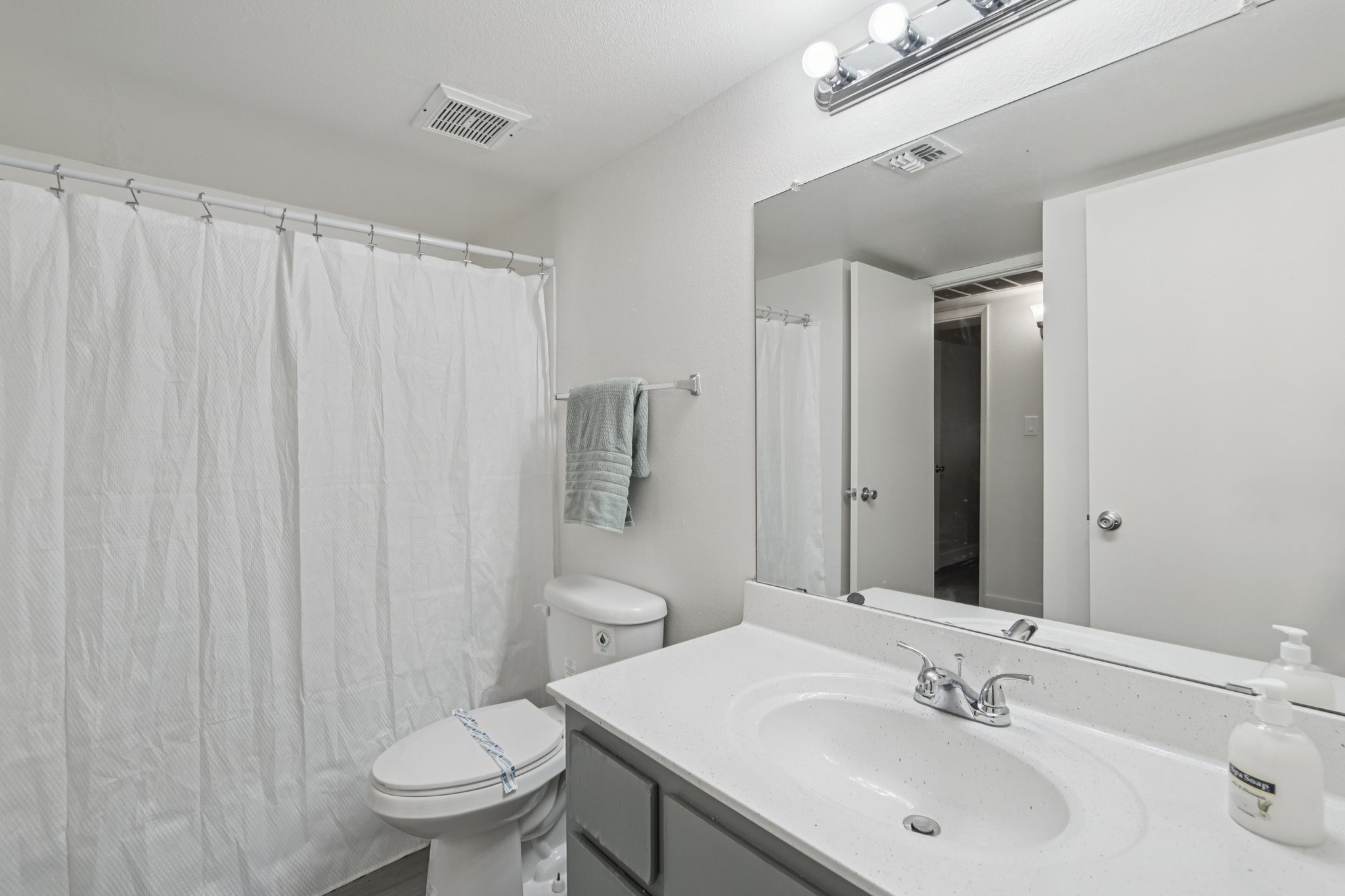 A clean, well-lit bathroom featuring a white shower curtain, a toilet, and a sink with a countertop. The mirror above the sink reflects the space, and a light fixture is installed above. A towel hangs on a rack, and there's a soap dispenser on the countertop. The decor is simple and modern.